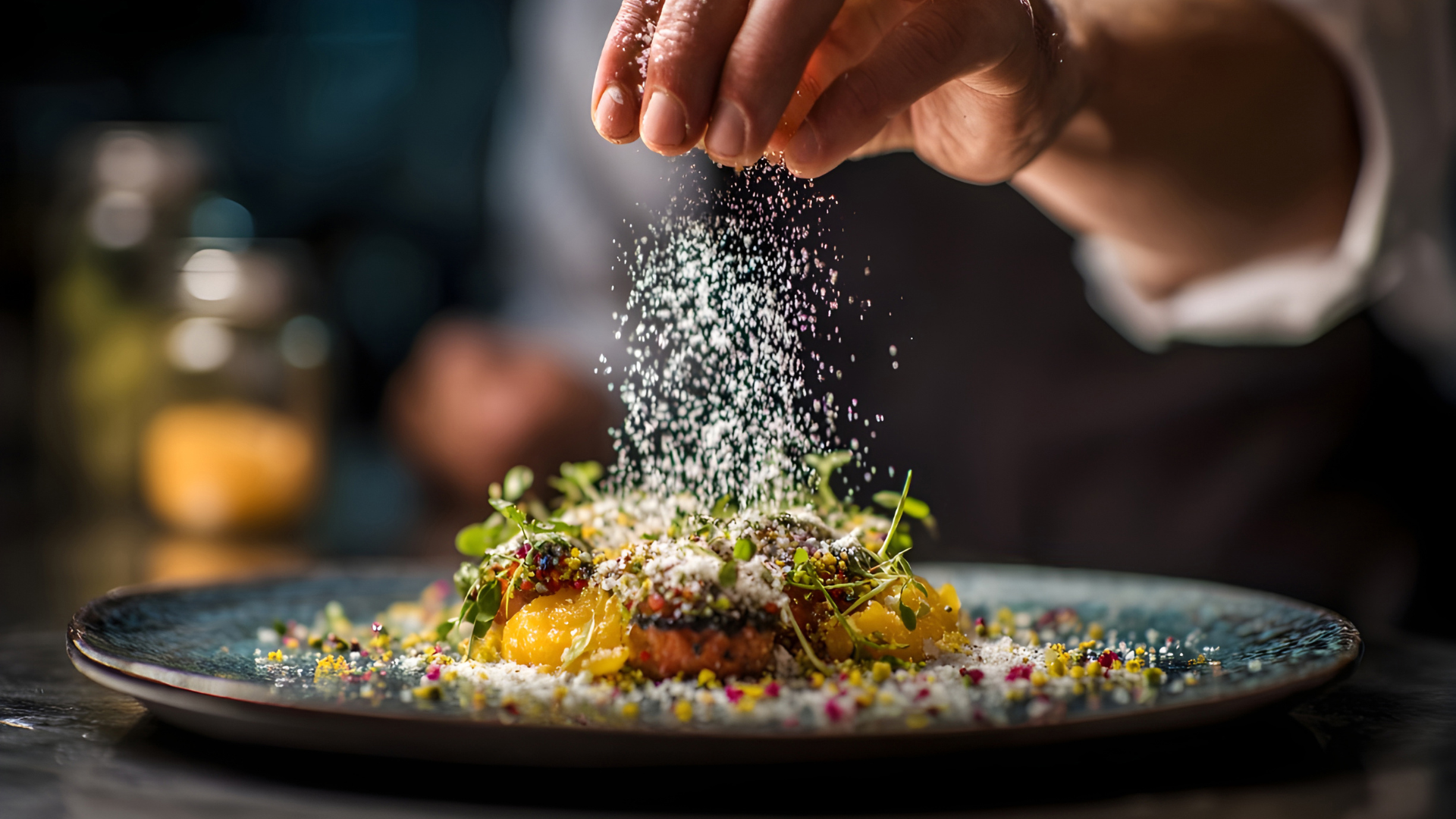 Uma mão de chef polvilha um ingrediente fino sobre um prato de alta cozinha cuidadosamente empratado, com flores, ervas e apontamentos coloridos, num ambiente escuro e desfocado que realça o detalhe e a precisão do gesto.