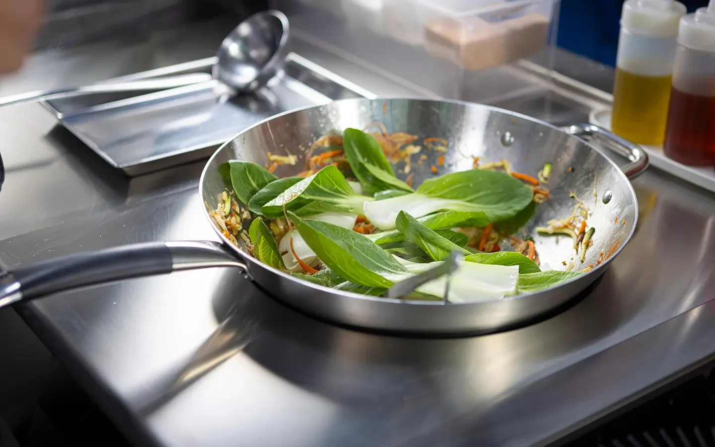 Wok sobre placa de indução numa cozinha profissional, com legumes salteados e folhas verdes frescas, junto a frascos de óleo e molho.