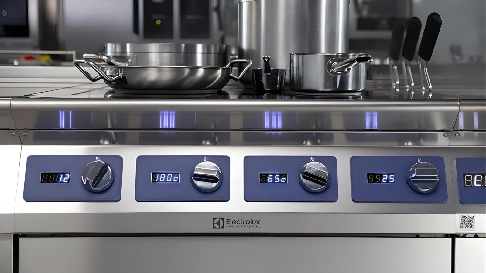 Linha de confeção profissional em inox com painéis de controlo e botões rotativos iluminados; tachos e utensílios sobre a superfície de trabalho numa cozinha industrial.