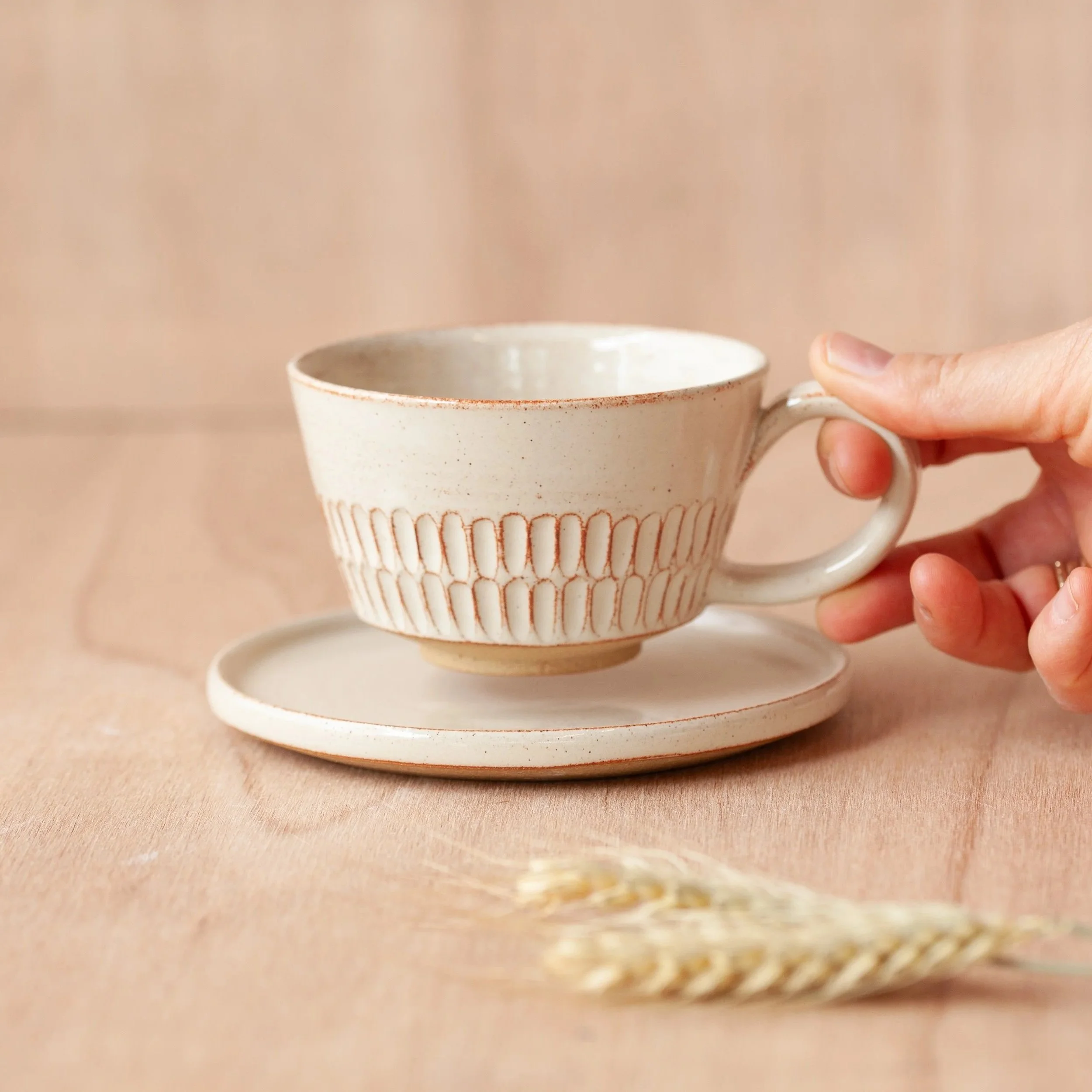 A person holding a ceramic mug with a textured ribbed design and a small handle, with a saucer underneath, against a neutral beige background.