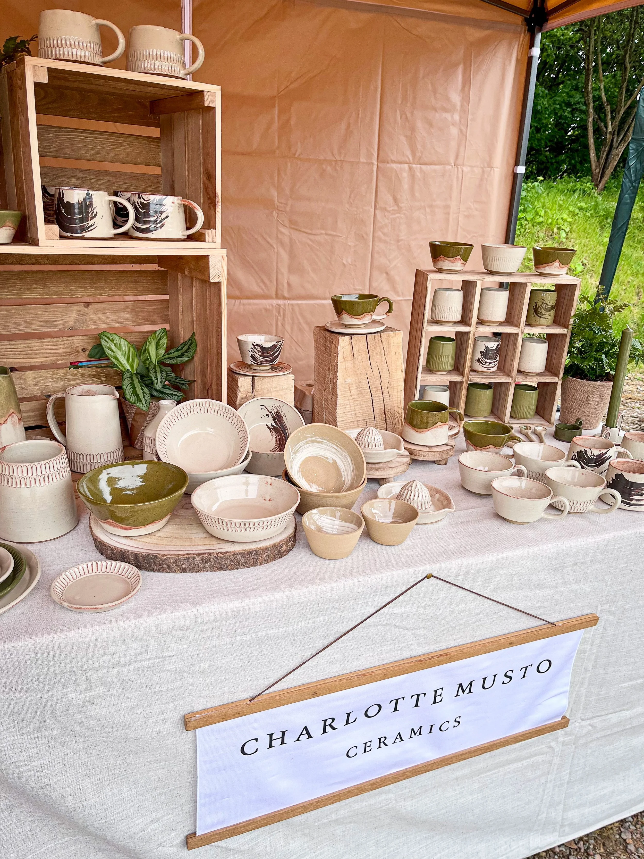 A display of handmade ceramic pottery including bowls, mugs, and plates in various earthy tones, arranged on wooden shelves and a table covered with a white cloth, labeled 'Charlottemusto Ceramics'.