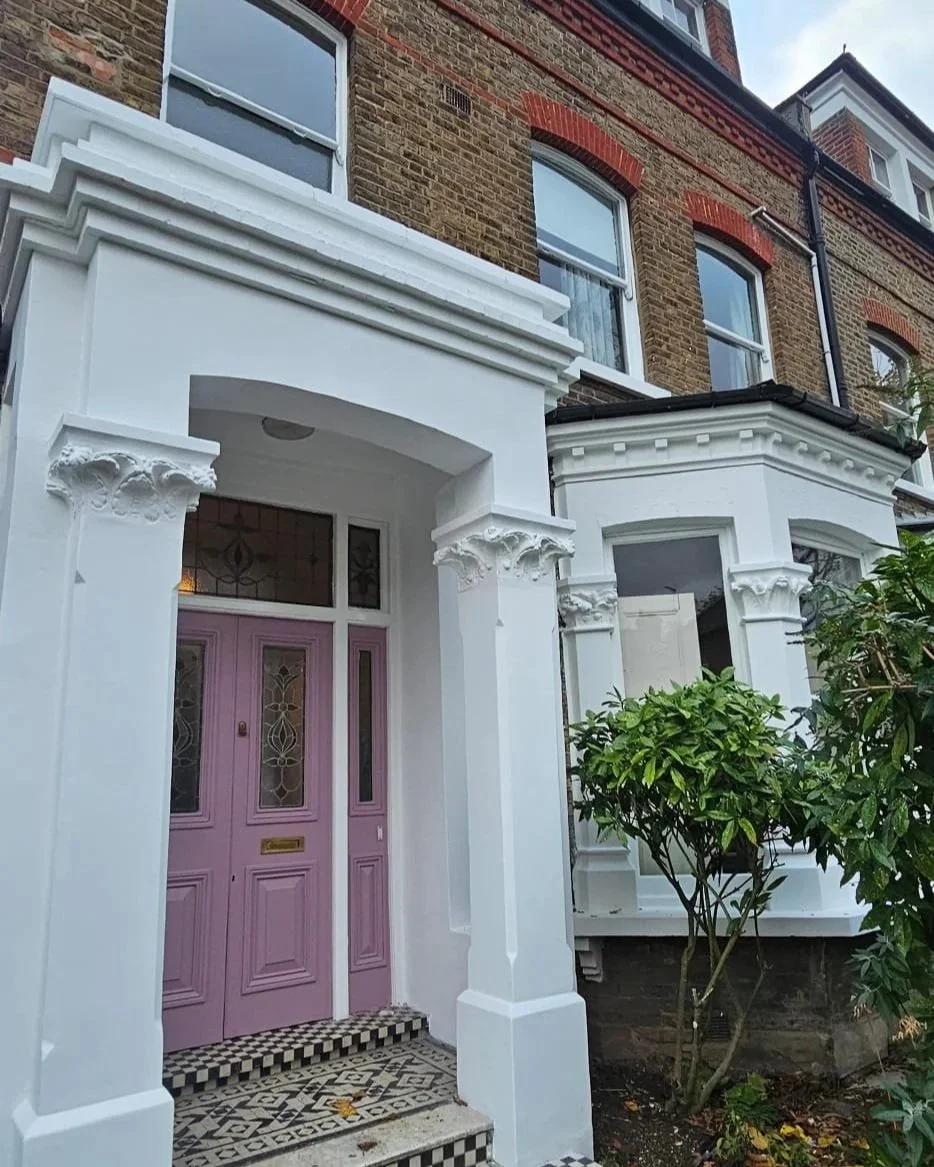 Close-up of a house entrance with pink double doors, decorative stained-glass windows, white ornate columns, and a patterned tile step, with greenery and brick walls in the background.