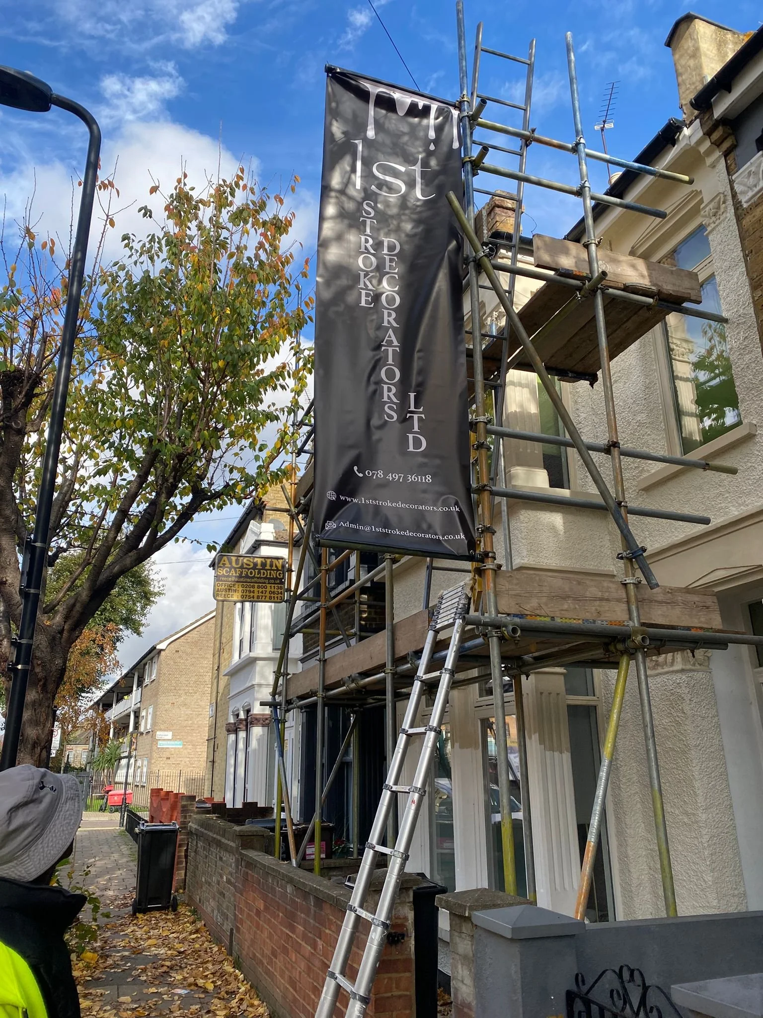 Building scaffolding with a vertical banner for 1st Stroke Decorators Ltd, on a street lined with trees and residential houses.