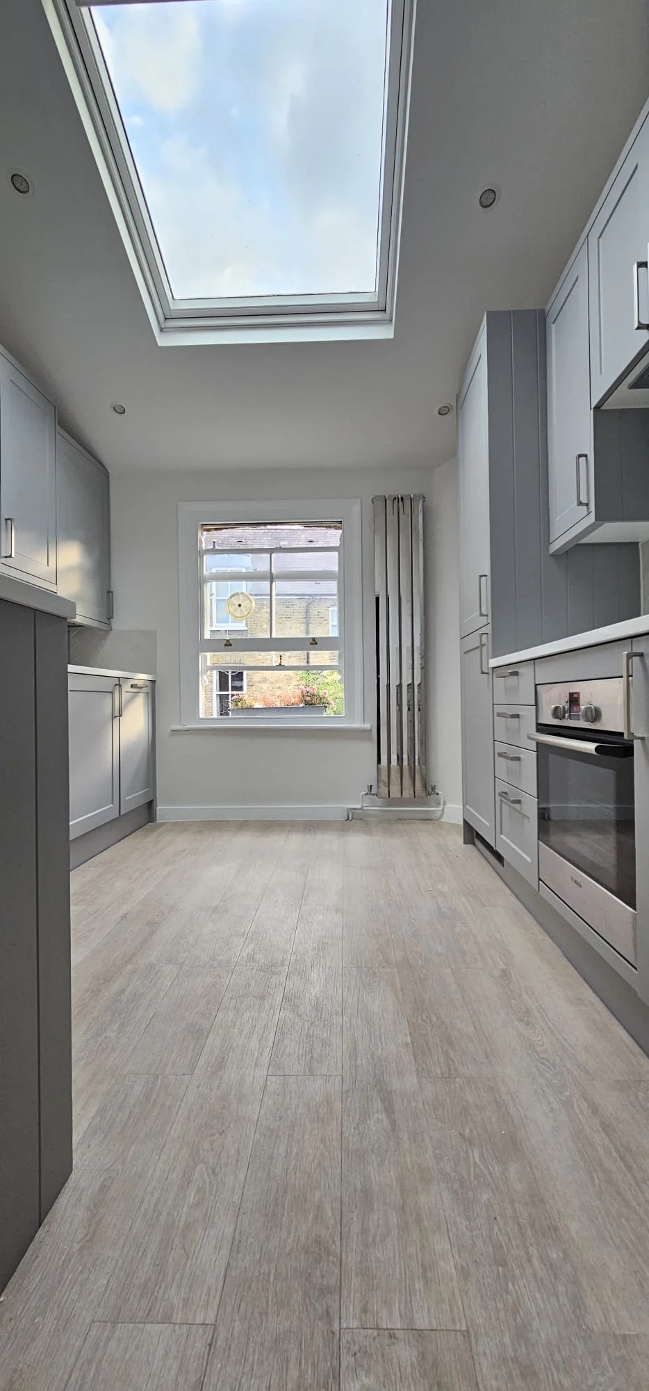 Empty kitchen with light wood flooring, white and gray cabinets, a large skylight, and a window showing part of a building and some greenery outside.