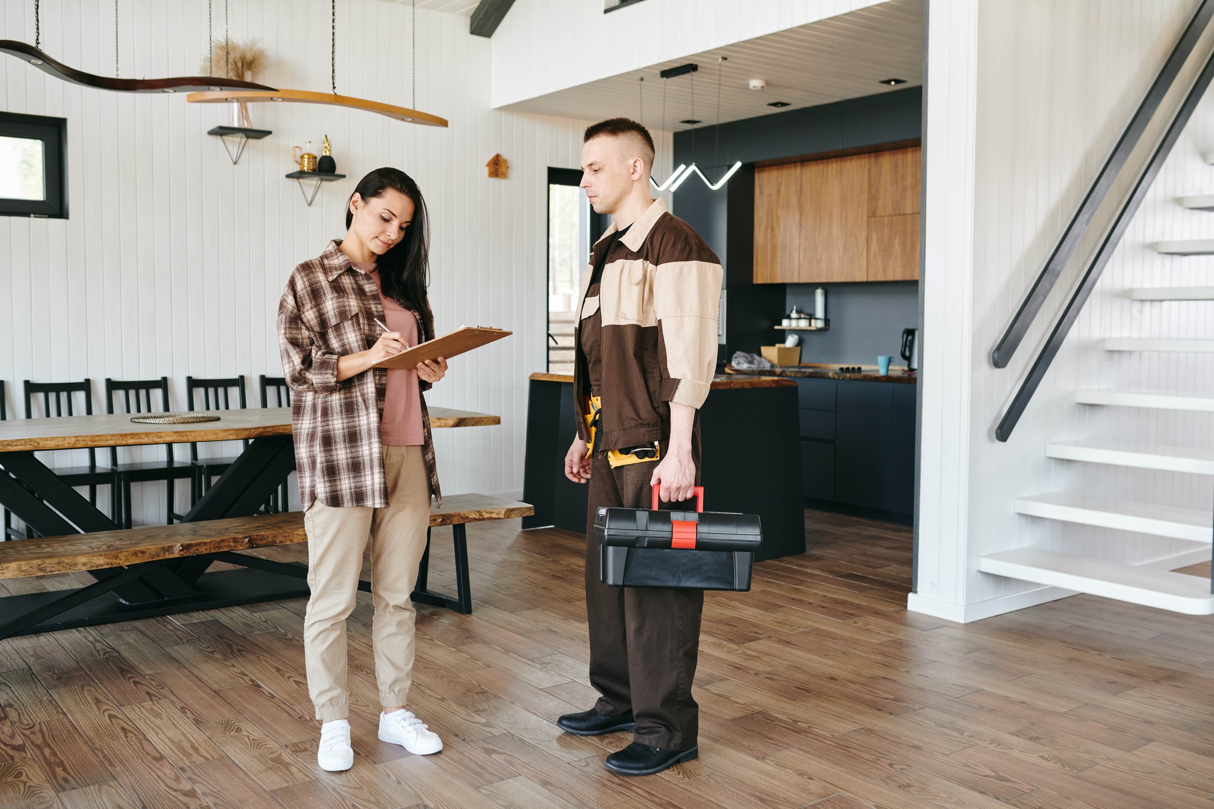 A woman in casual clothes writing on a clipboard engaged in conversation with a man in work attire holding a toolbox, inside a modern kitchen and dining area.