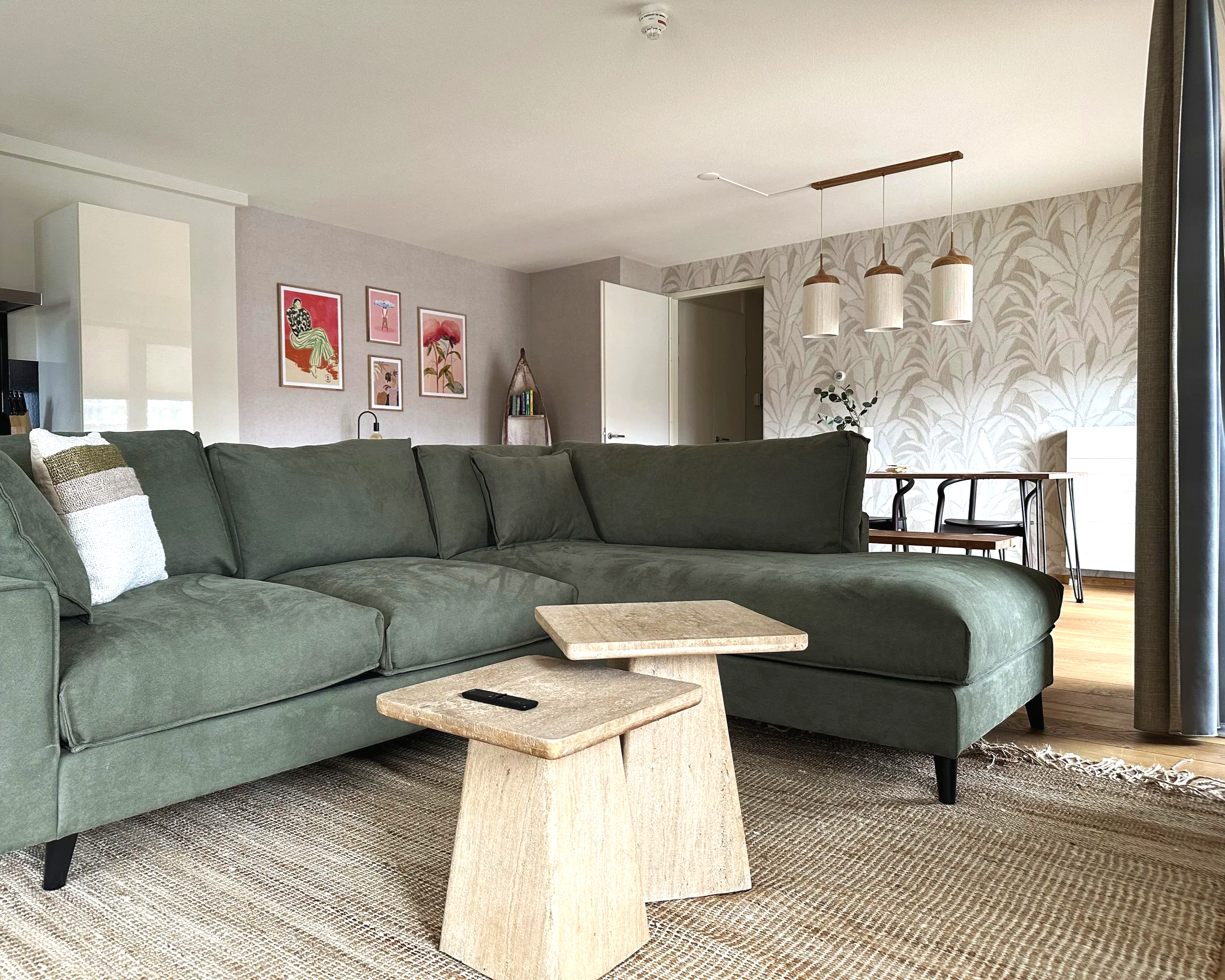 A living room with a large green sectional sofa, a beige coffee table, a patterned area rug, wall art, and a dining area with a pendant light fixture.