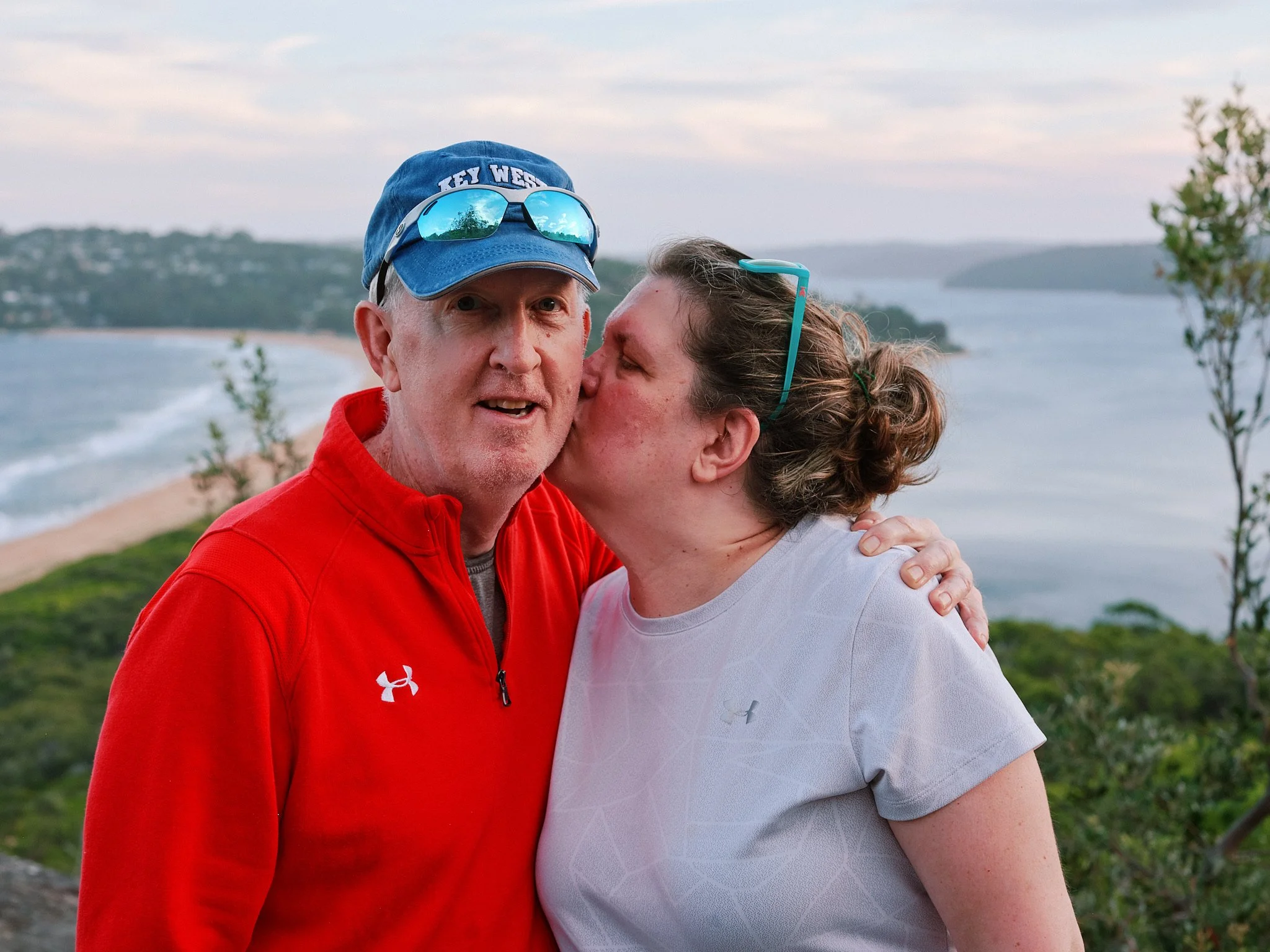 A woman kisses a man on the cheek outdoors with a scenic view of a beach, water, and distant hills.