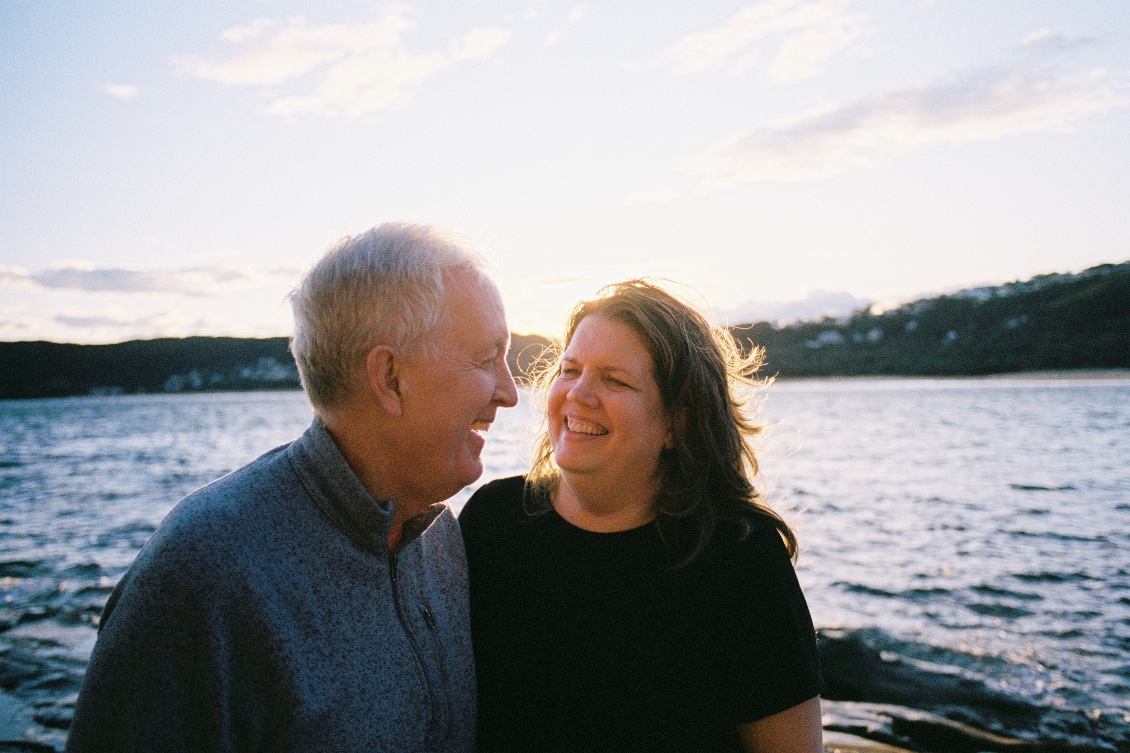 A joyful elderly man and woman sharing a happy moment by a lake during sunset.