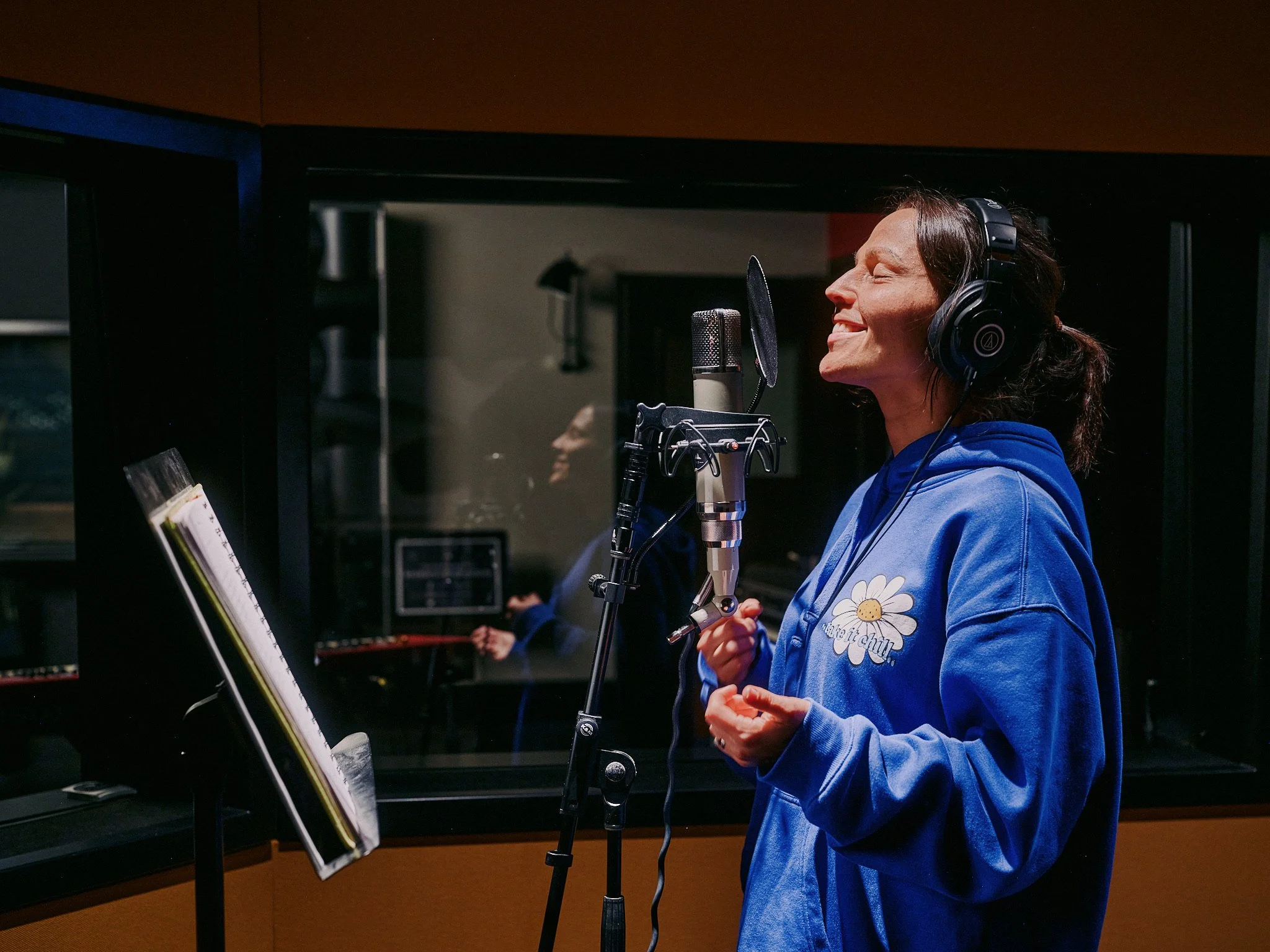 Woman in a blue hoodie singing passionately in a recording studio, wearing headphones, in front of a microphone.