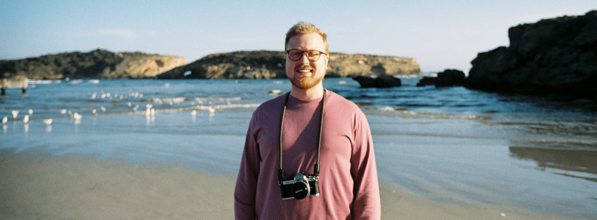 A man with glasses and a beard smiling at the camera on a beach, wearing a pink shirt, with a camera hanging from his neck, and seagulls flying in the background near the water and cliffs.