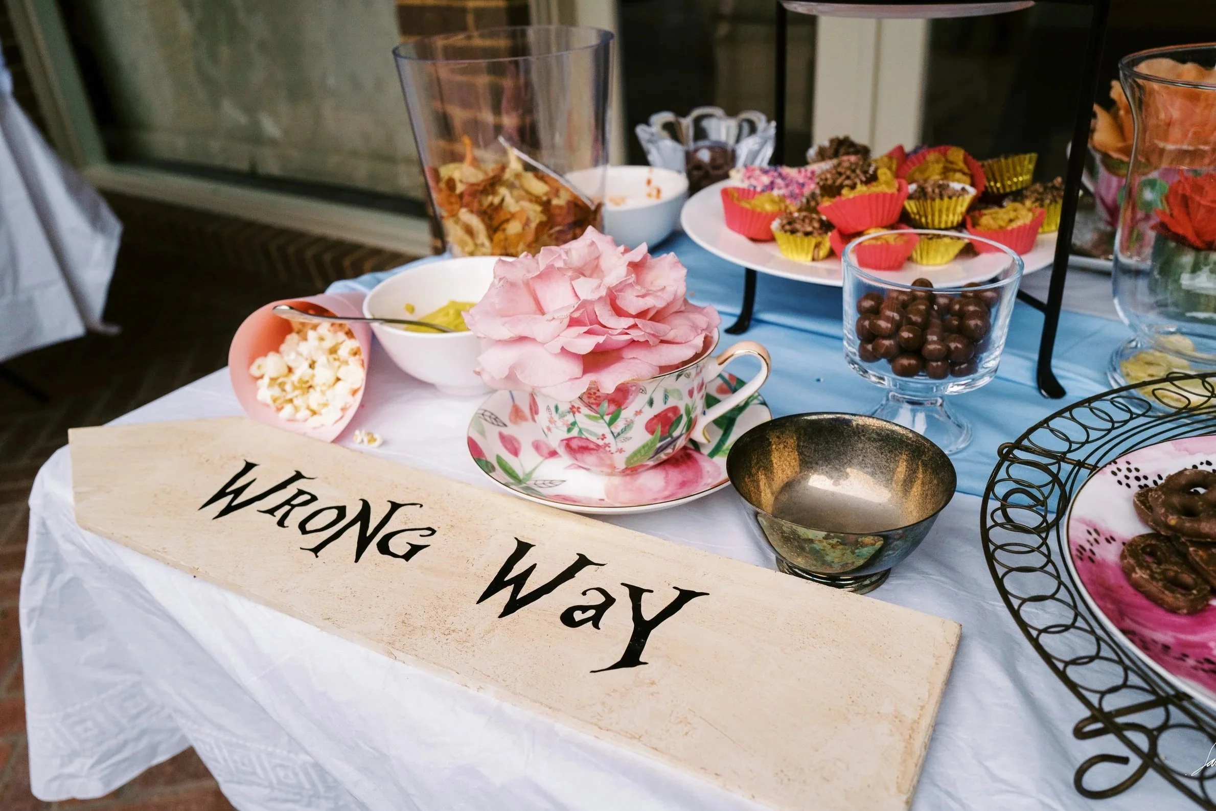 Colorful snack table with pink and floral teacup, bowls of popcorn, chocolates, and cupcakes, featuring a wooden sign that reads 'WRONG WAY'.