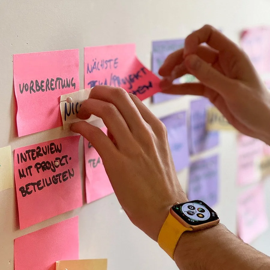 Person, die rosafarbene und lilafarbene Haftnotizen mit handgeschriebenem deutschen Text an einer Wand anbringt und eine gelbe Smartwatch trägt.