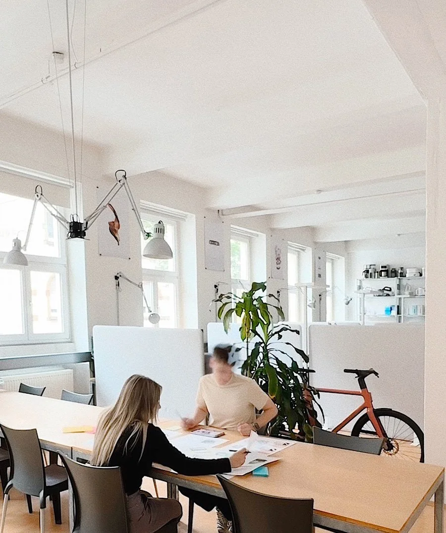 Zwei Mitarbeiter sitzen an einem Konferenztisch aus hellem Holz in einem hellen, modernen Büro, mit einem Fahrrad und einer großen Pflanze im Hintergrund.
