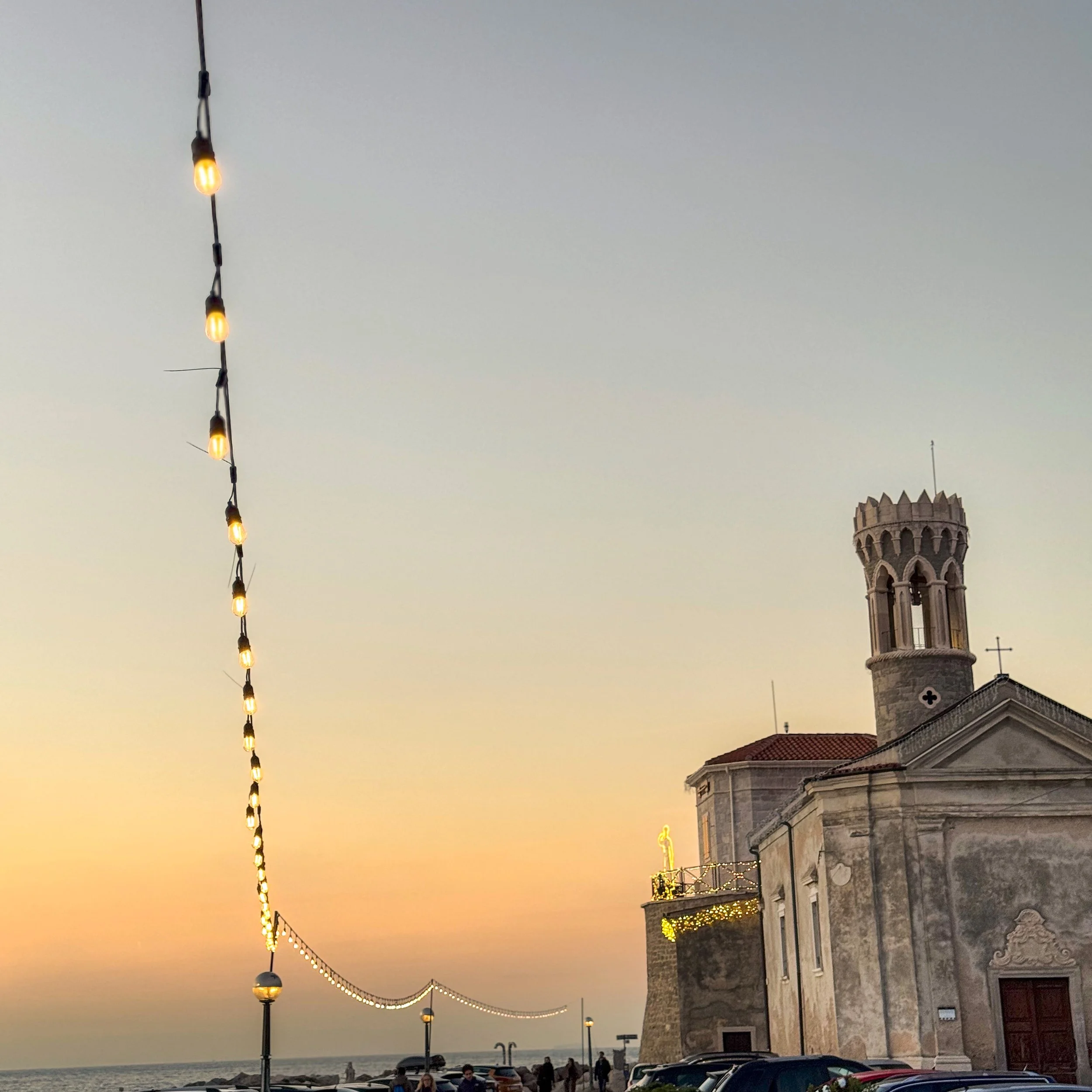 Golden hour sunset in Piran coastal town with illuminated church, string lights, and guests strolling near luxury transfer vehicles