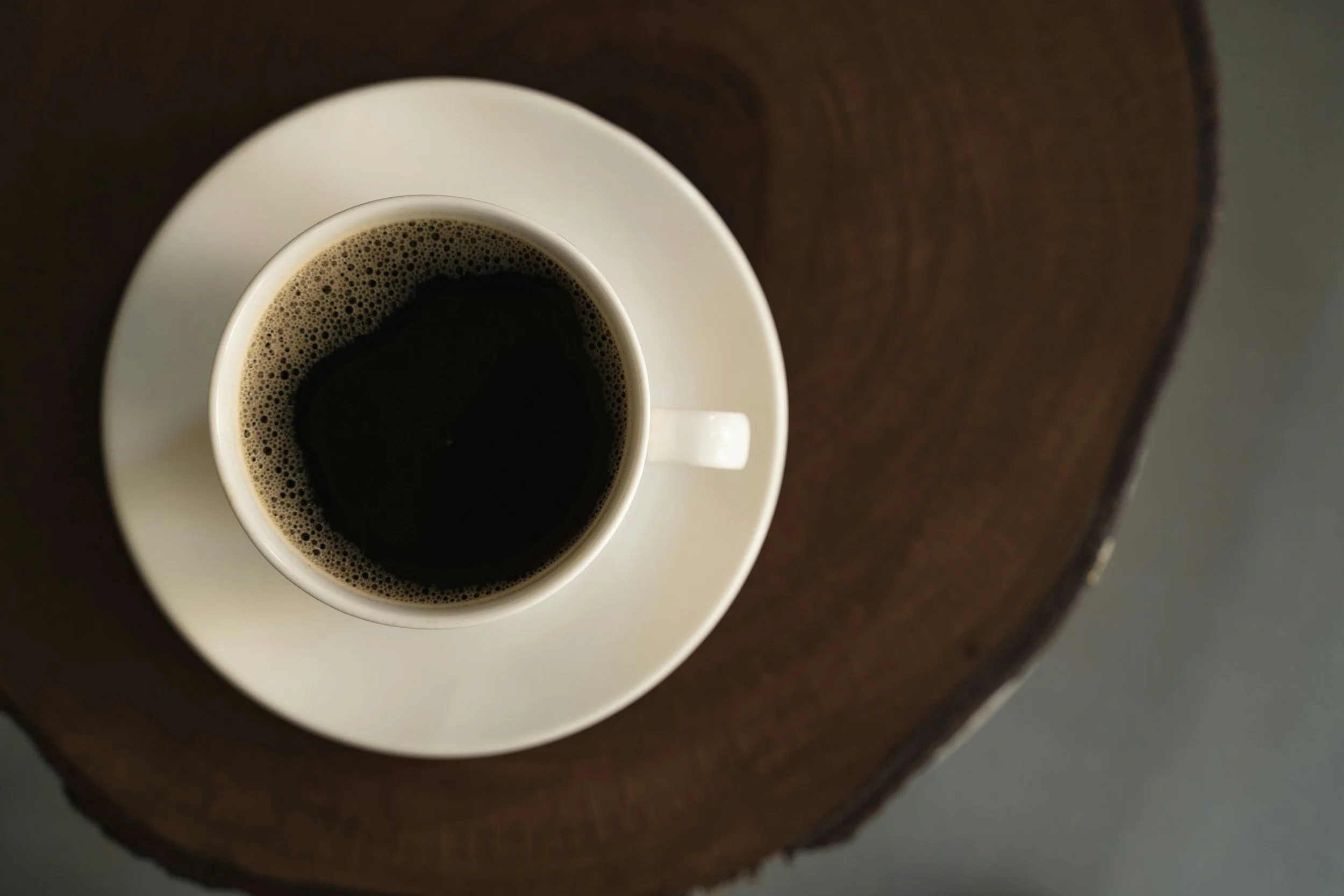 A white coffee cup filled with black coffee sitting on a matching white saucer, viewed from above, placed on a circular wooden table.