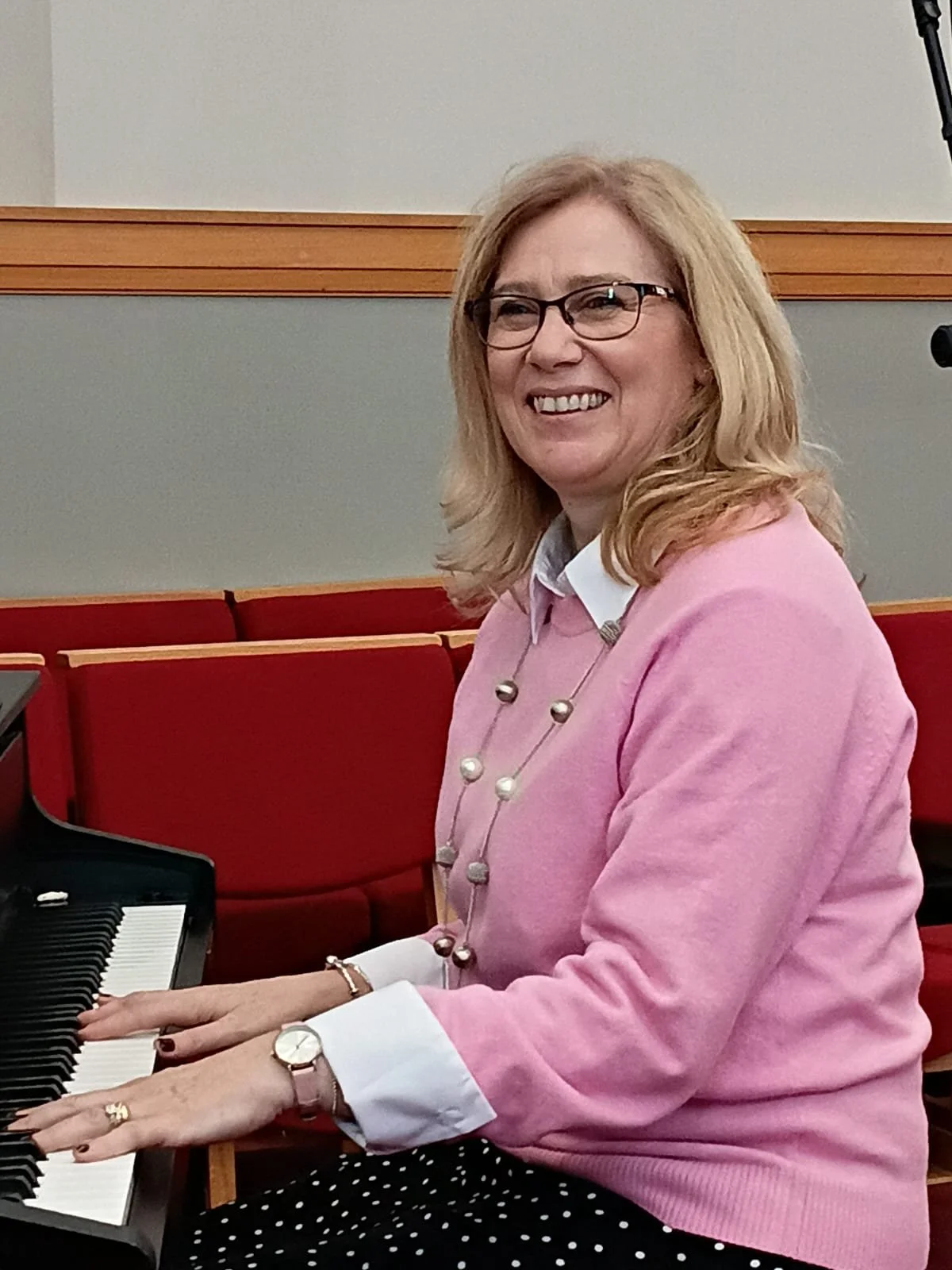A smiling woman with glasses, blonde hair, and wearing a pink jumper over a white shirt, and a necklace sitting at a piano.