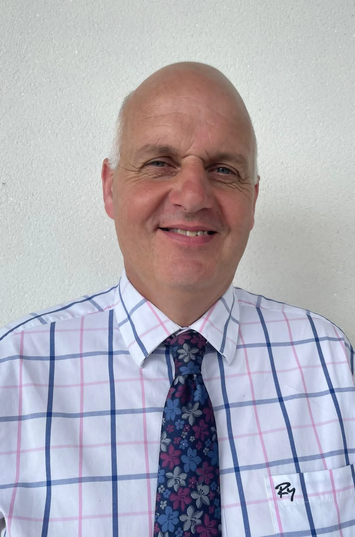 A smiling middle-aged man with a bald head in a white shirt with plaid pattern, wearing a floral tie, standing against a plain white wall.