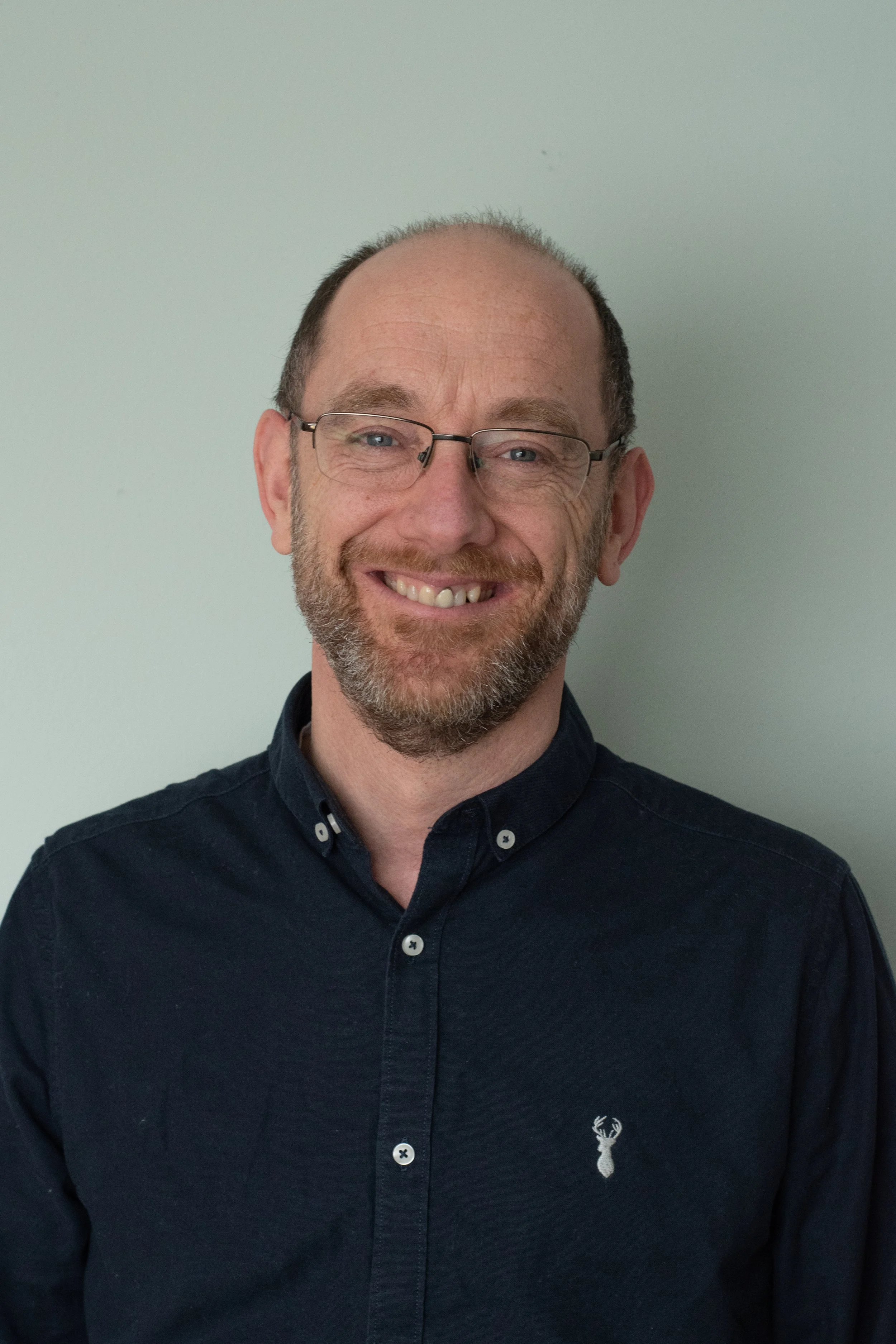 A man with glasses and a beard smiling, wearing a black button-up shirt standing against a plain light-colored wall.