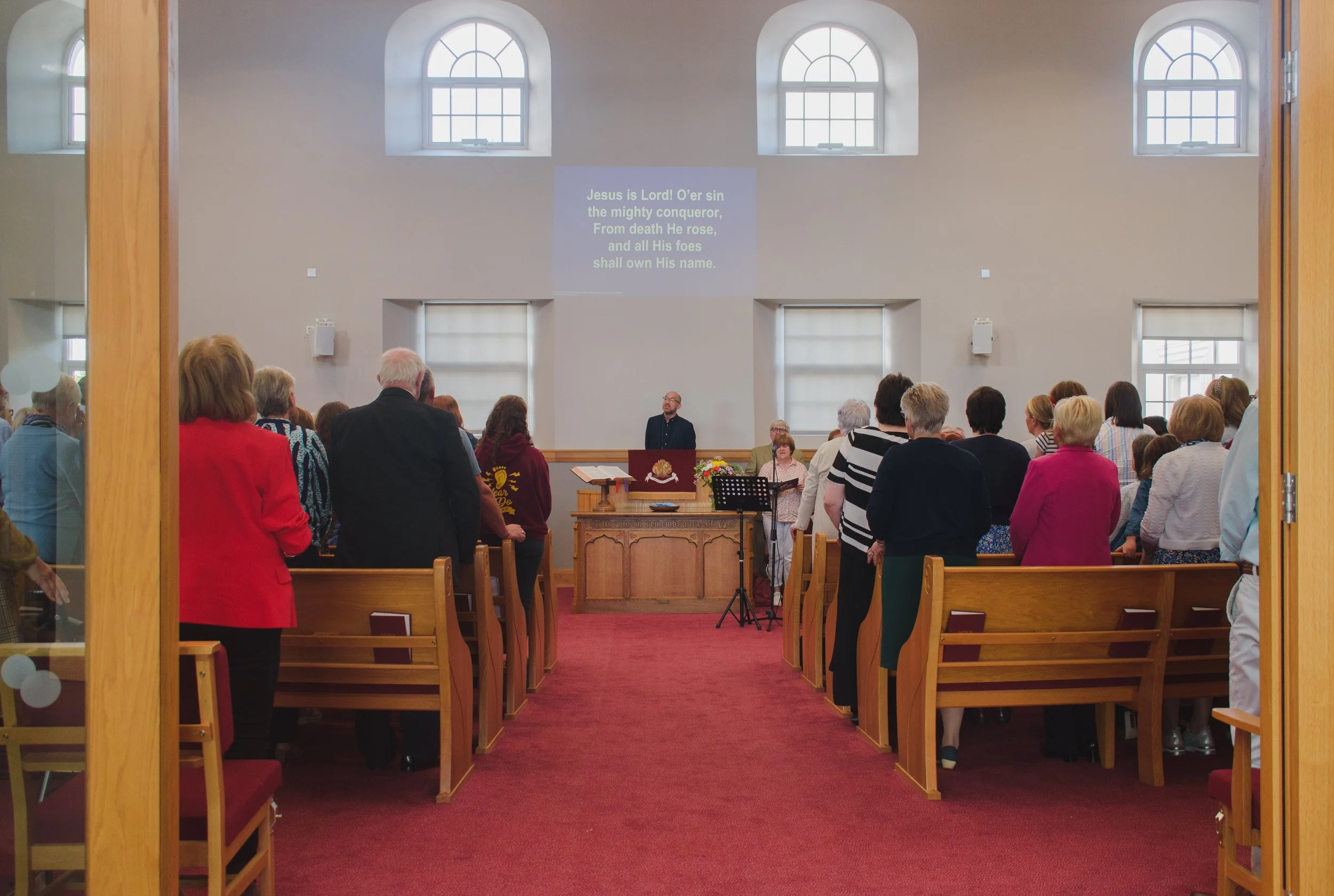 A group of people gathered in a church, standing and facing the pulpit during a service. The church has white walls, arched windows, and a red carpet. A preacher is at the pulpit, and a song or hymn with lyrics projected on the wall behind him.