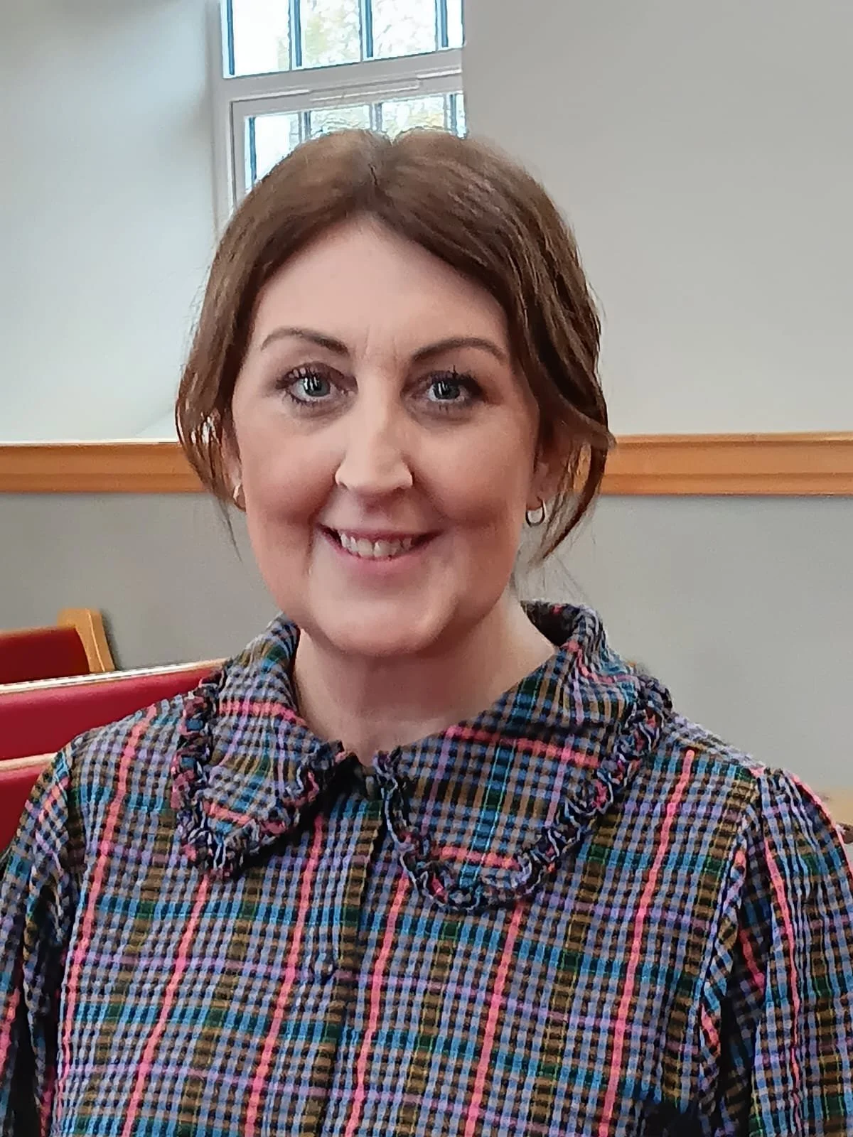A smiling woman with short brown hair and blue eyes, wearing a colorful plaid shirt, sitting in a room with a window and red chairs.