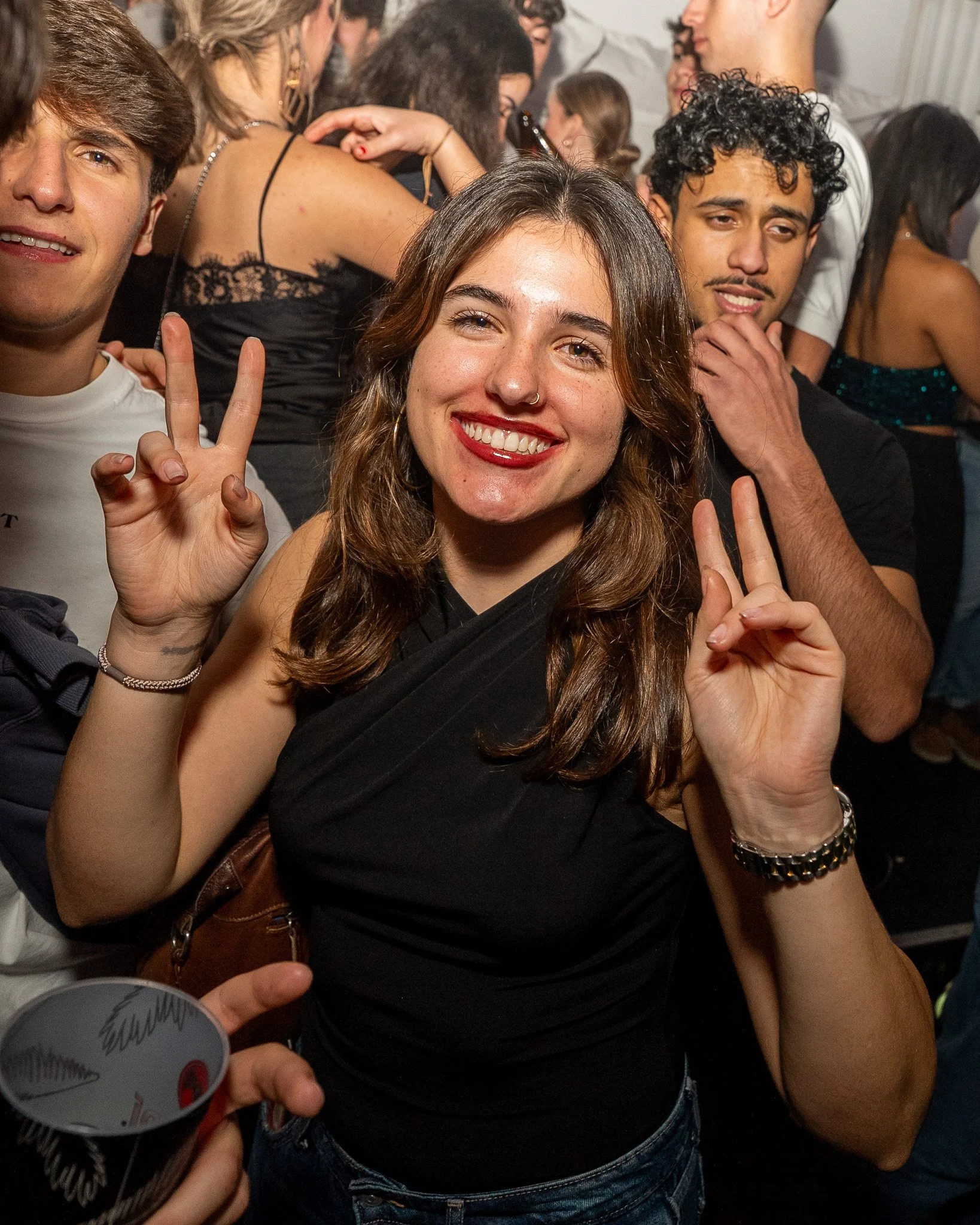 Young woman smiling and making peace signs with both hands at a party or social gathering with people in the background.