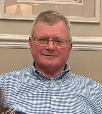 A middle-aged man with glasses wearing a blue checkered shirt, sitting indoors.