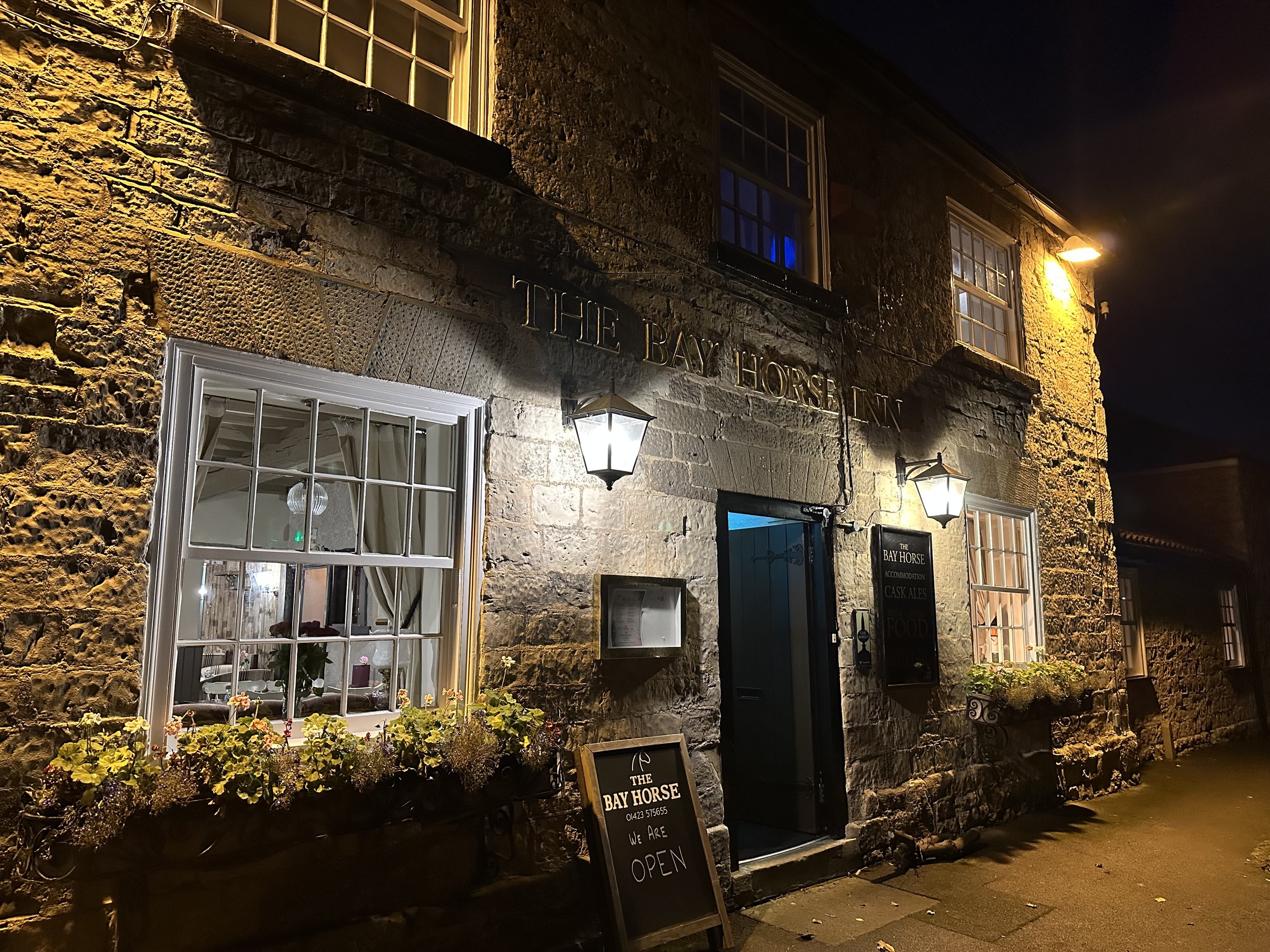 The exterior of a rustic stone building with illuminated signs and window boxes of flowers, nighttime scene.