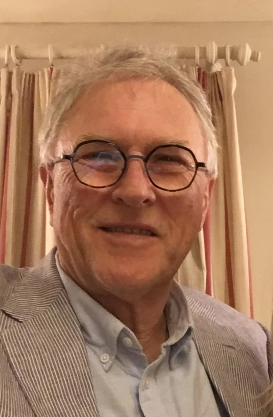 A smiling man with gray hair, round glasses, wearing a striped blazer and a light-colored dress shirt, standing in front of beige and red curtains.