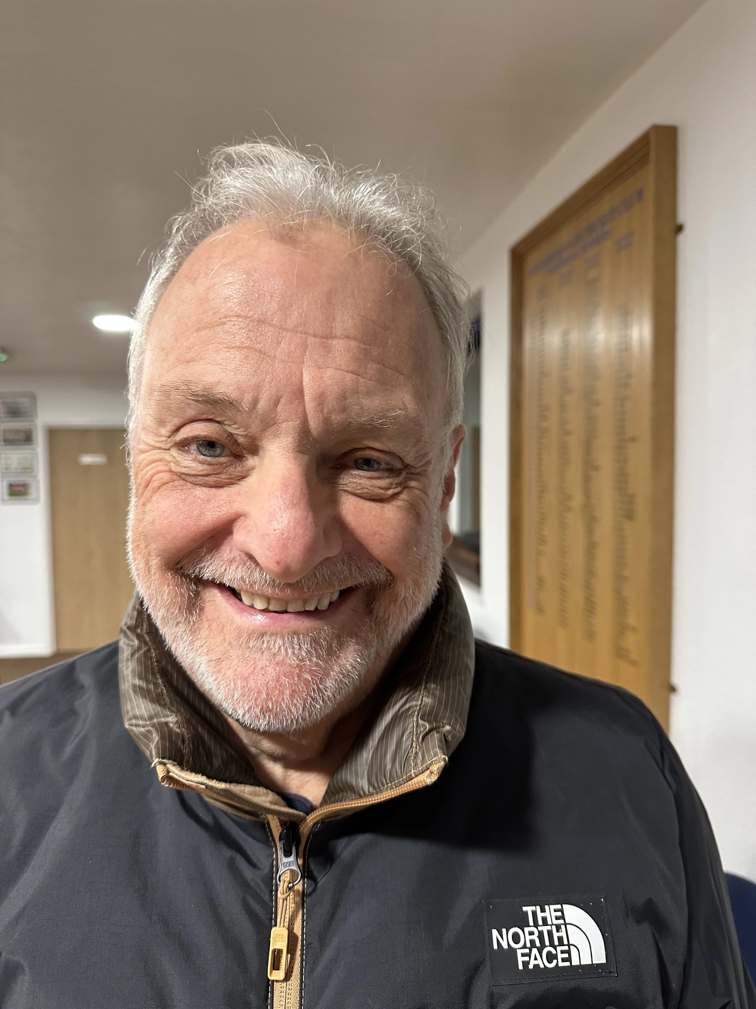 Close-up of a smiling older man with gray hair and beard, wearing a black The North Face jacket, inside a building with light-colored walls and a wooden plaque in the background.