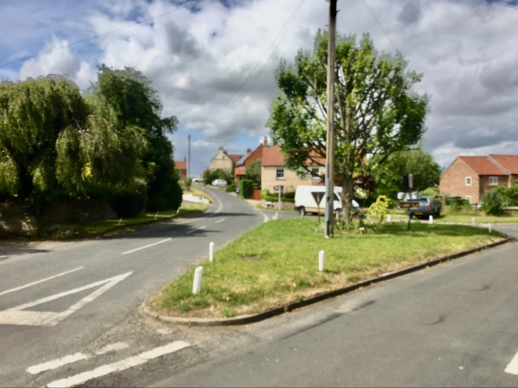 A suburban street with a small traffic island with grass and a tree, surrounded by houses, cars, and utility poles, under a partly cloudy sky.