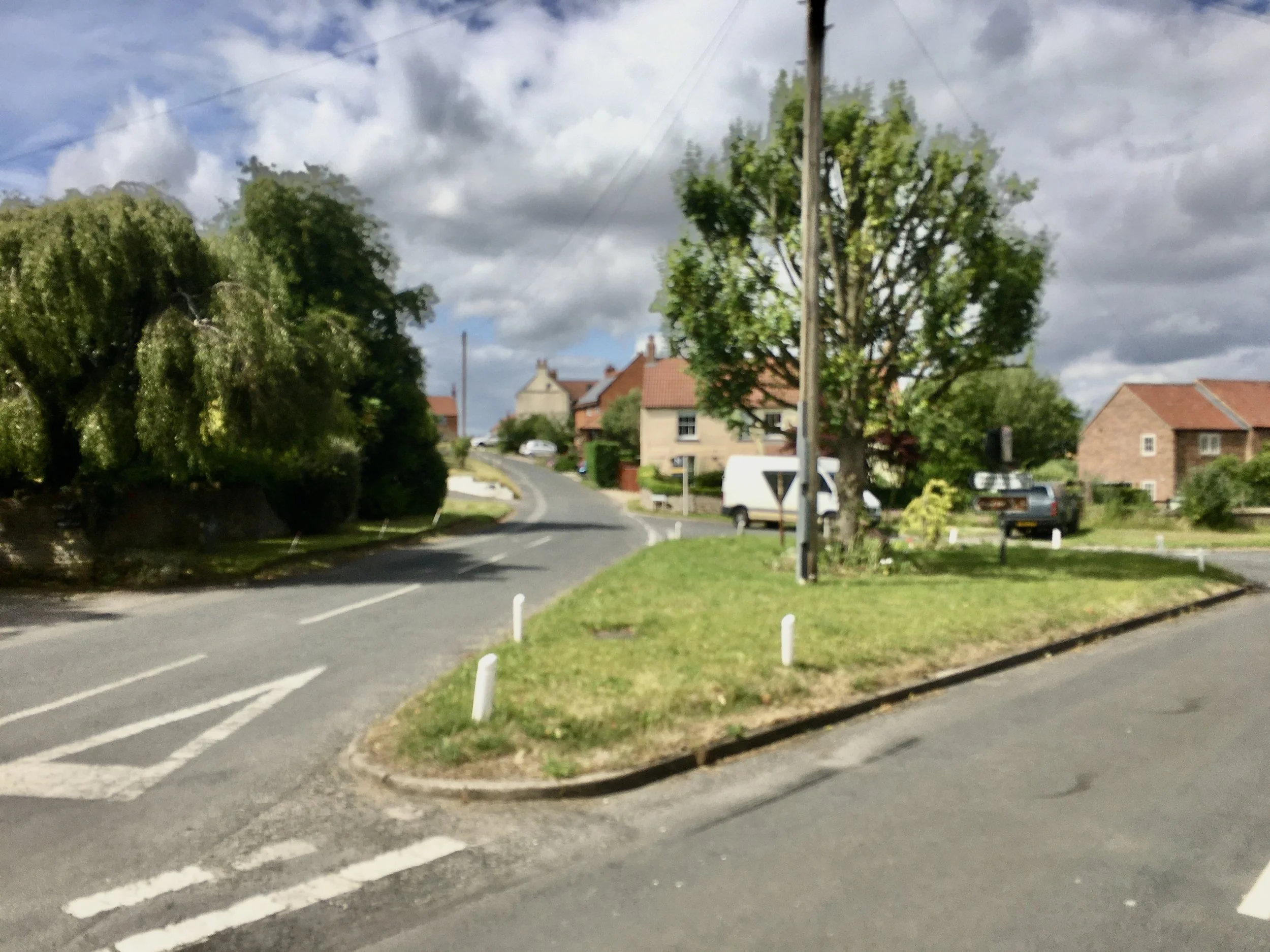 A small residential street with houses, green trees, a white van, and parked cars, under a partly cloudy sky.