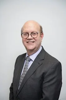 A middle-aged man with glasses, wearing a gray suit and tie, smiling against a plain light background.