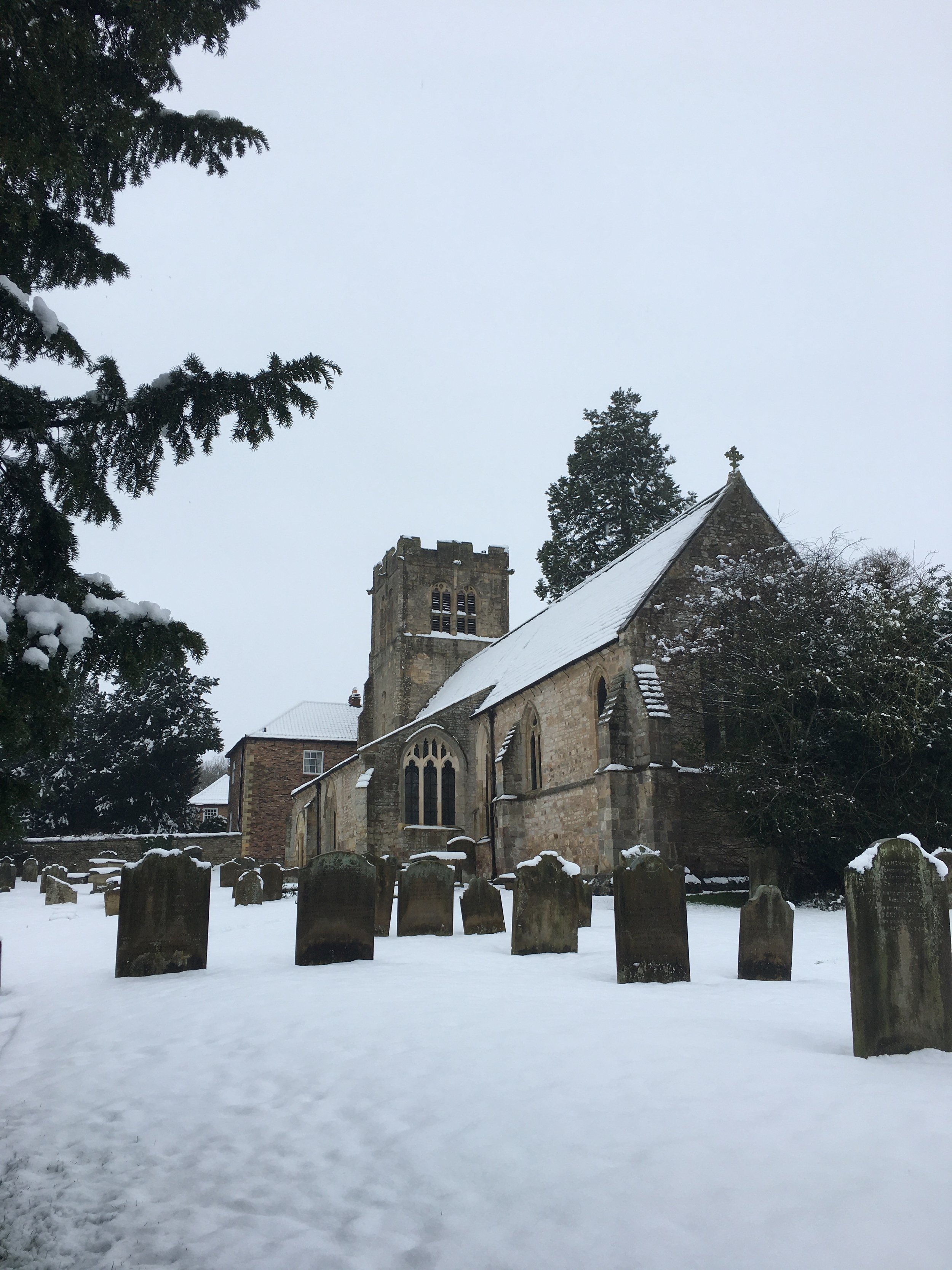 Snow-covered church with cemetery in foreground, with gravestones and trees surrounding the church.