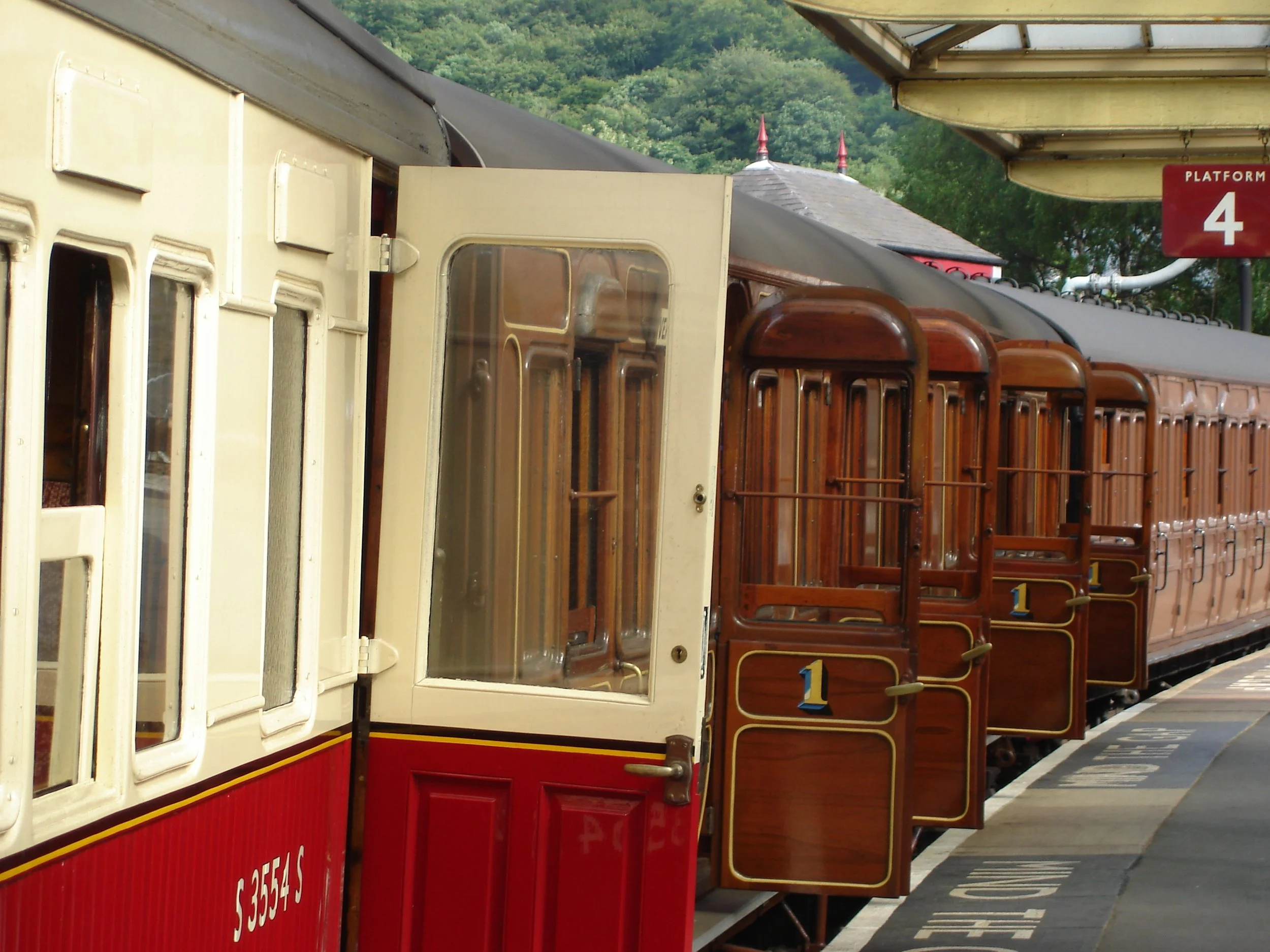 A vintage train at a station platform with multiple wooden and painted cars, a red platform sign indicating platform 4, and green hills in the background.