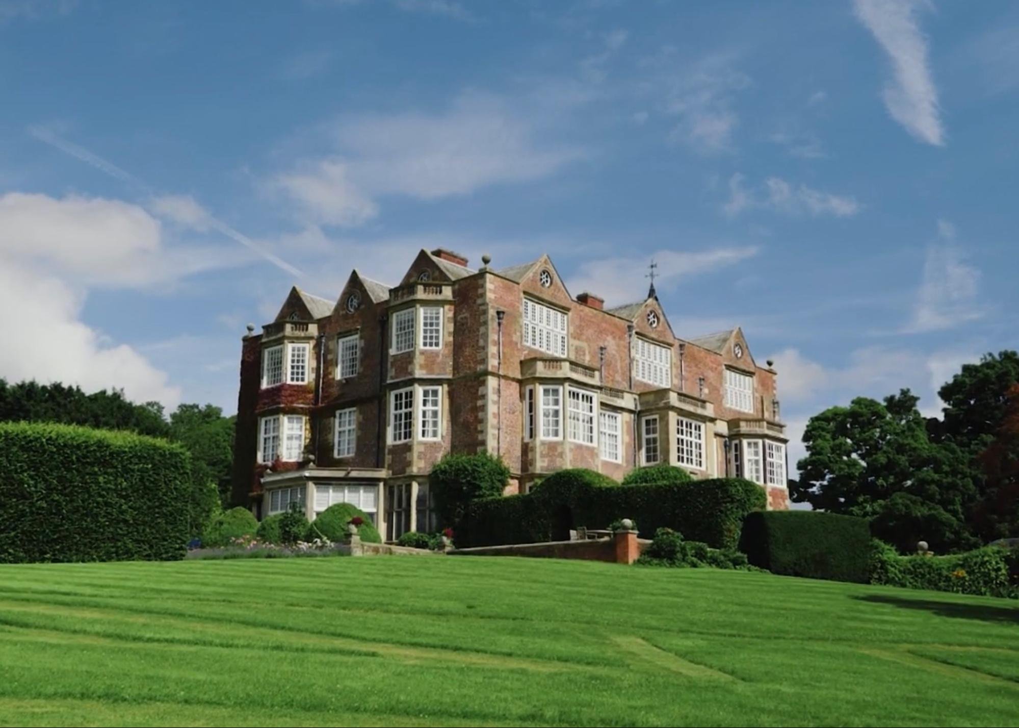A large, historic brick mansion with numerous windows, green landscaping, and a well-manicured lawn under a blue sky.