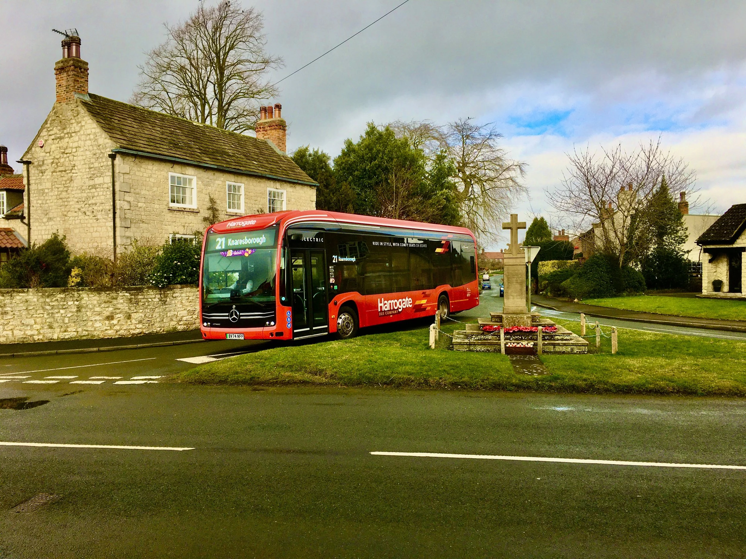 A red bus with HTML 21 on its display parked near a roadside memorial with a stone cross and flower arrangement, in a small town with houses and trees under a cloudy sky.