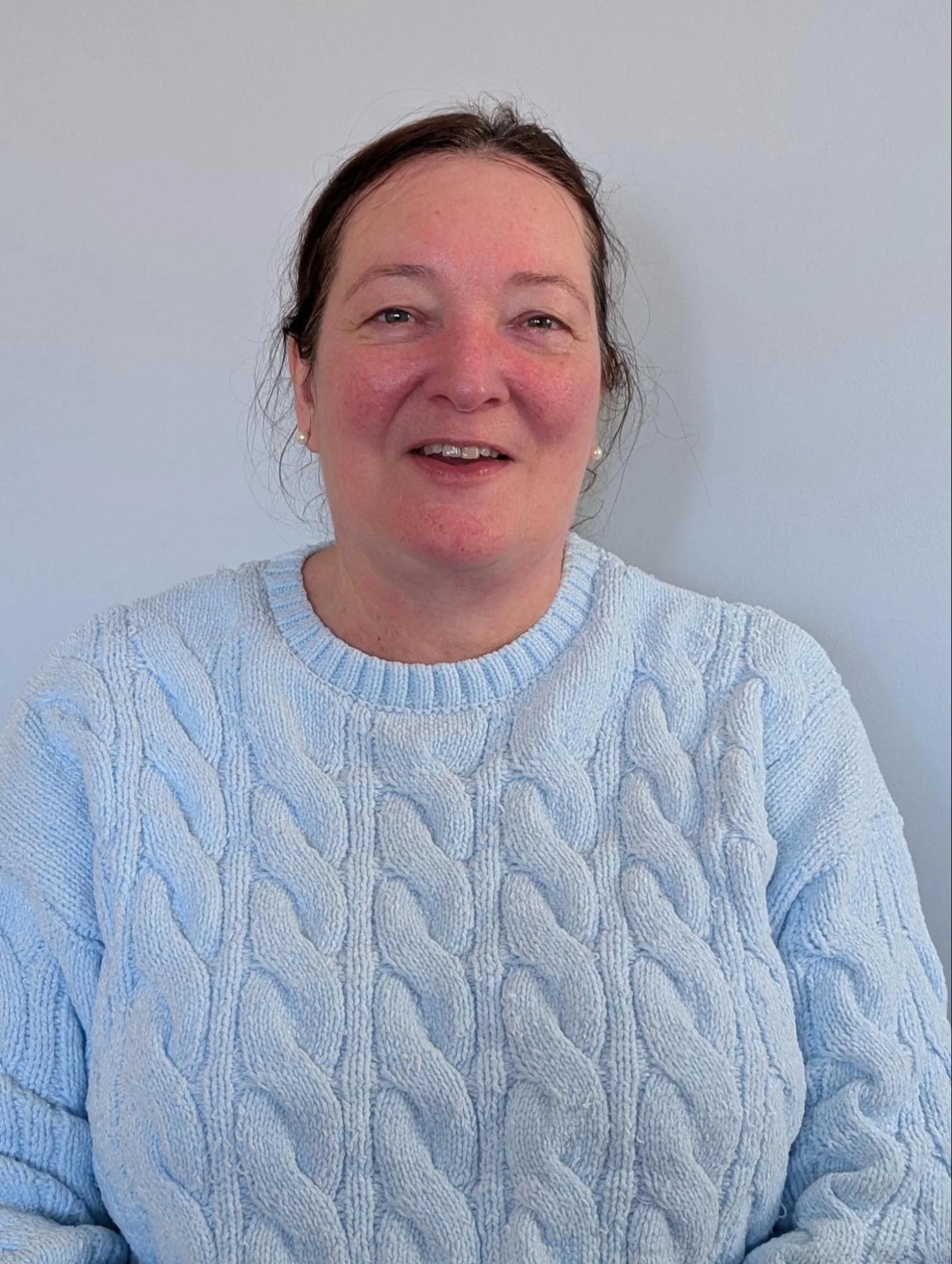 A woman with brown hair wearing a light blue cable knit sweater, smiling at the camera with a plain light-colored wall background.