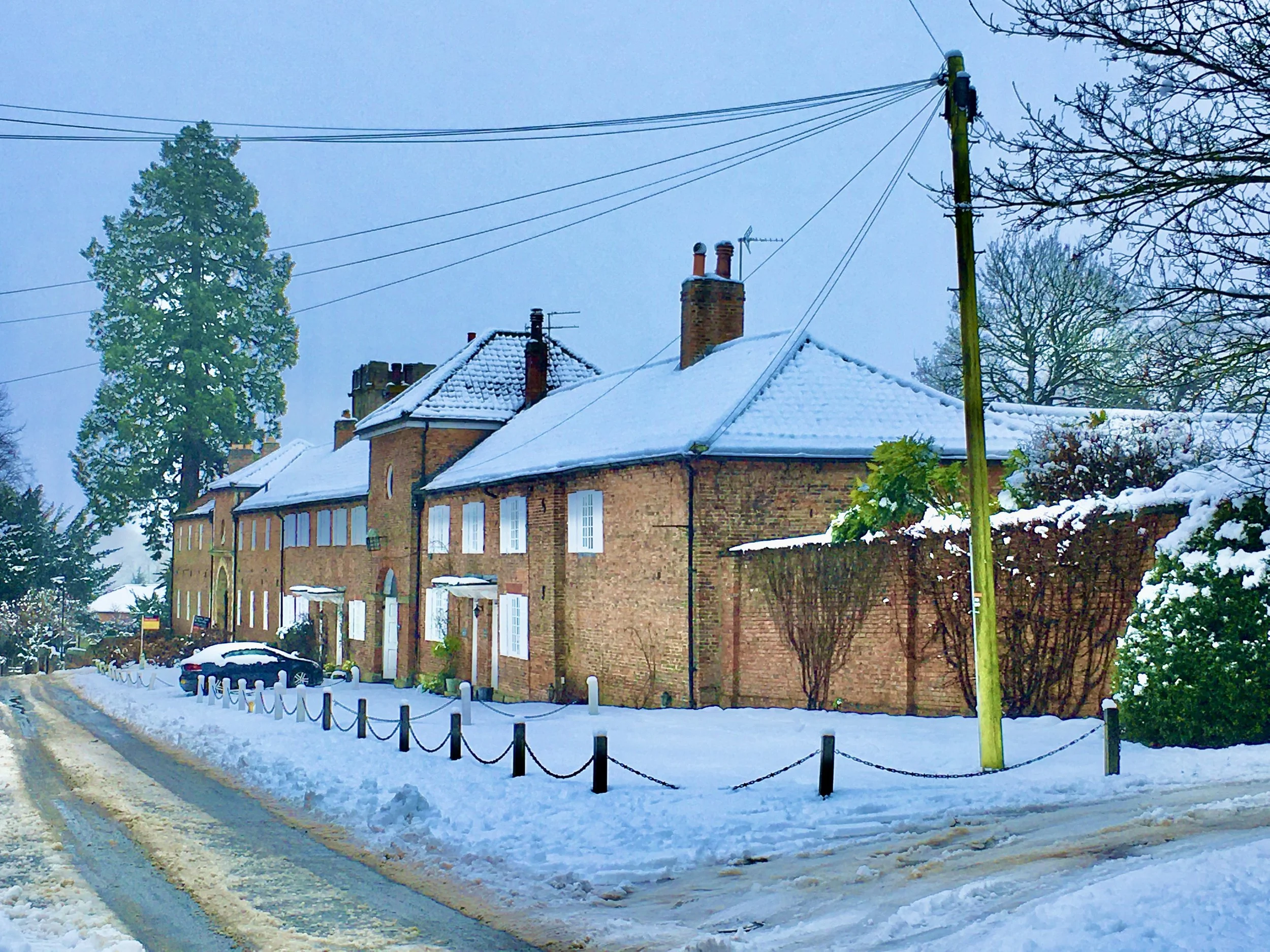 Snow-covered brick buildings with white window frames across a snow-covered street with tire tracks, a black car, and a snow-covered sidewalk enclosed by a black post-and-chain fence, utility poles, leafless trees, and cloudy sky.