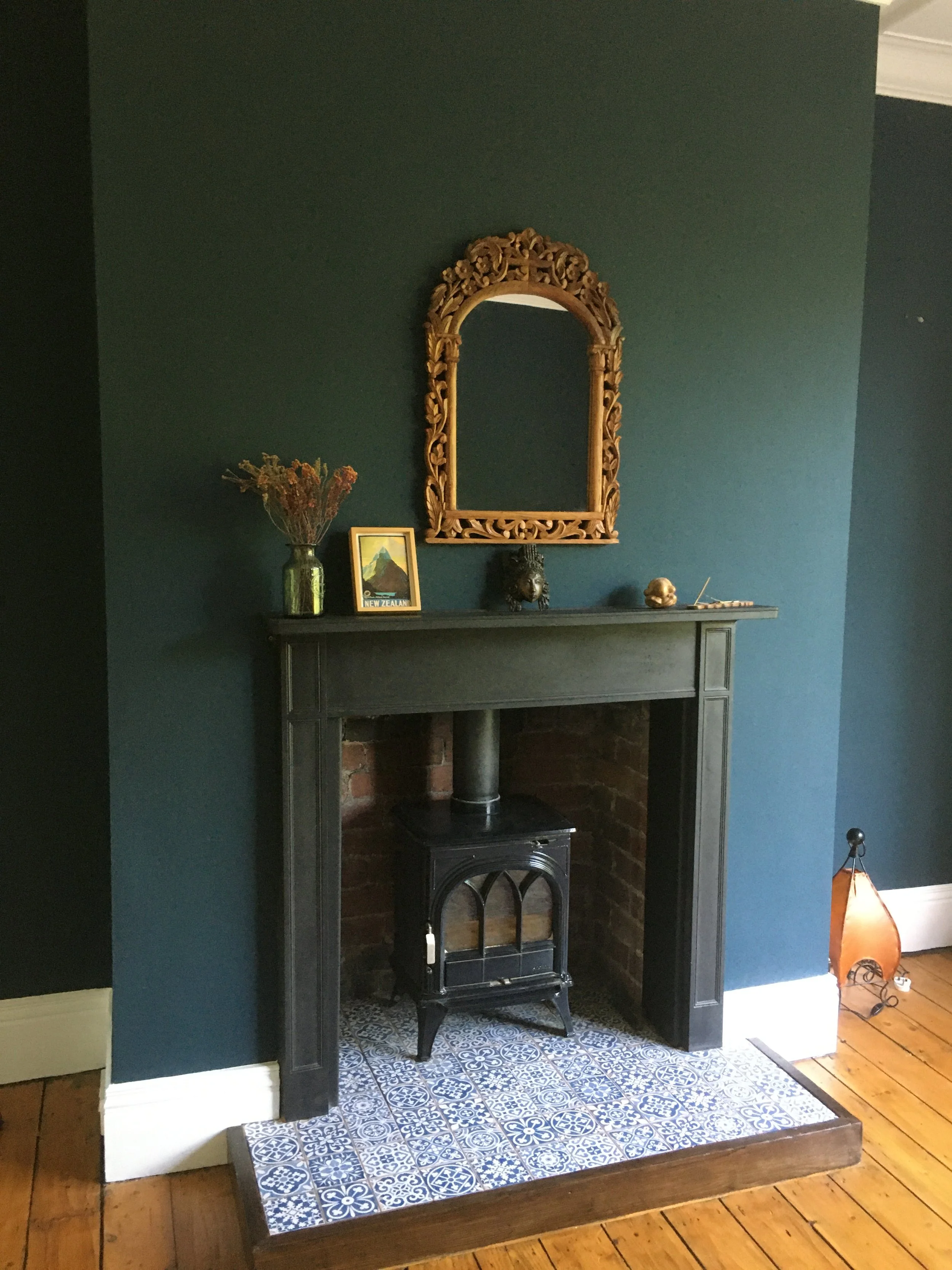 A black fireplace with a wood stove inside, set on a decorative tile hearth, with a dark green wall and a mirror above, decorated with a vase of dried flowers, a framed postcard, a small sculpture, and miscellaneous items.