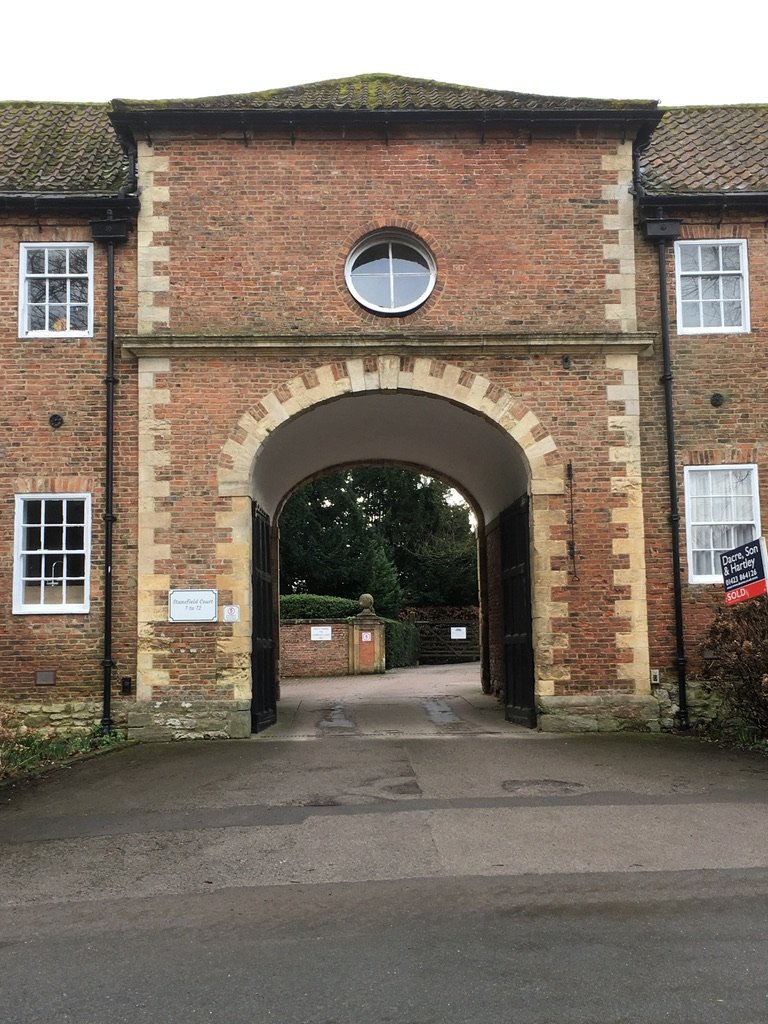 Brick building with an arched gateway and a round window above it, leading to a courtyard. Windows on either side of the gateway, some with curtains, and a sign indicating the property is sold.