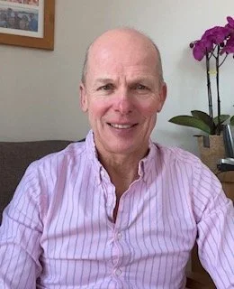 A smiling older man in a pink striped shirt sitting in a living room next to a purple orchid plant.