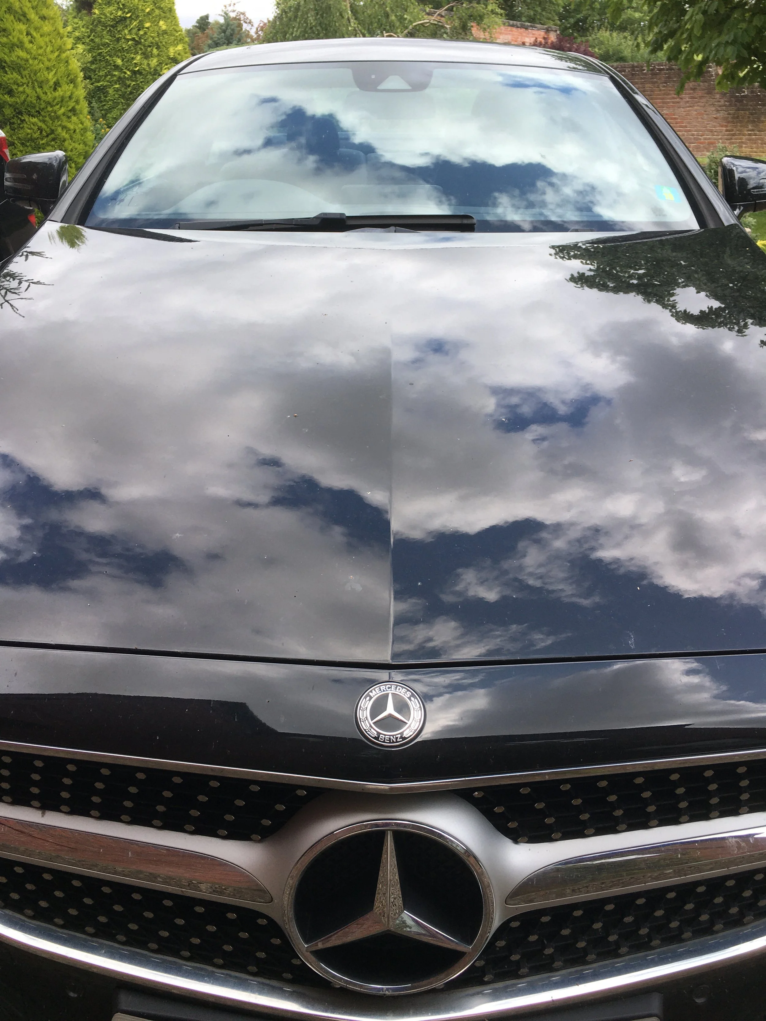 Front view of a black Mercedes-Benz car with its hood reflecting cloudy sky and trees.