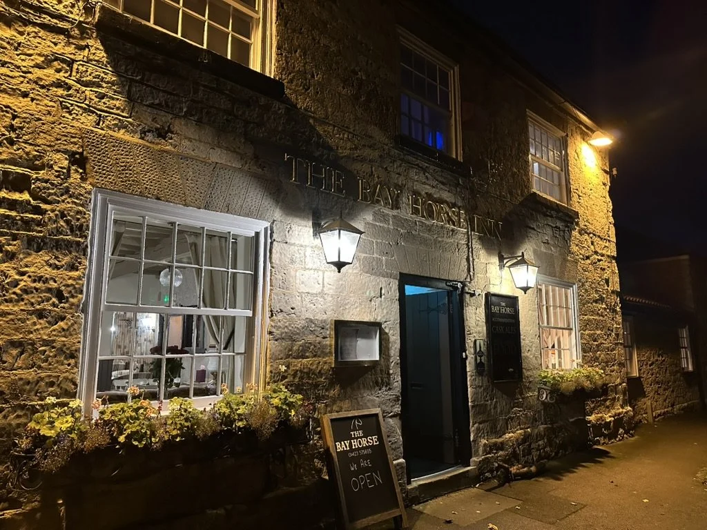 Exterior view of The Bay Horse Inn, a stone building with lit lanterns, windows with flower boxes, and a chalkboard sign outside reading 'Open' at night.