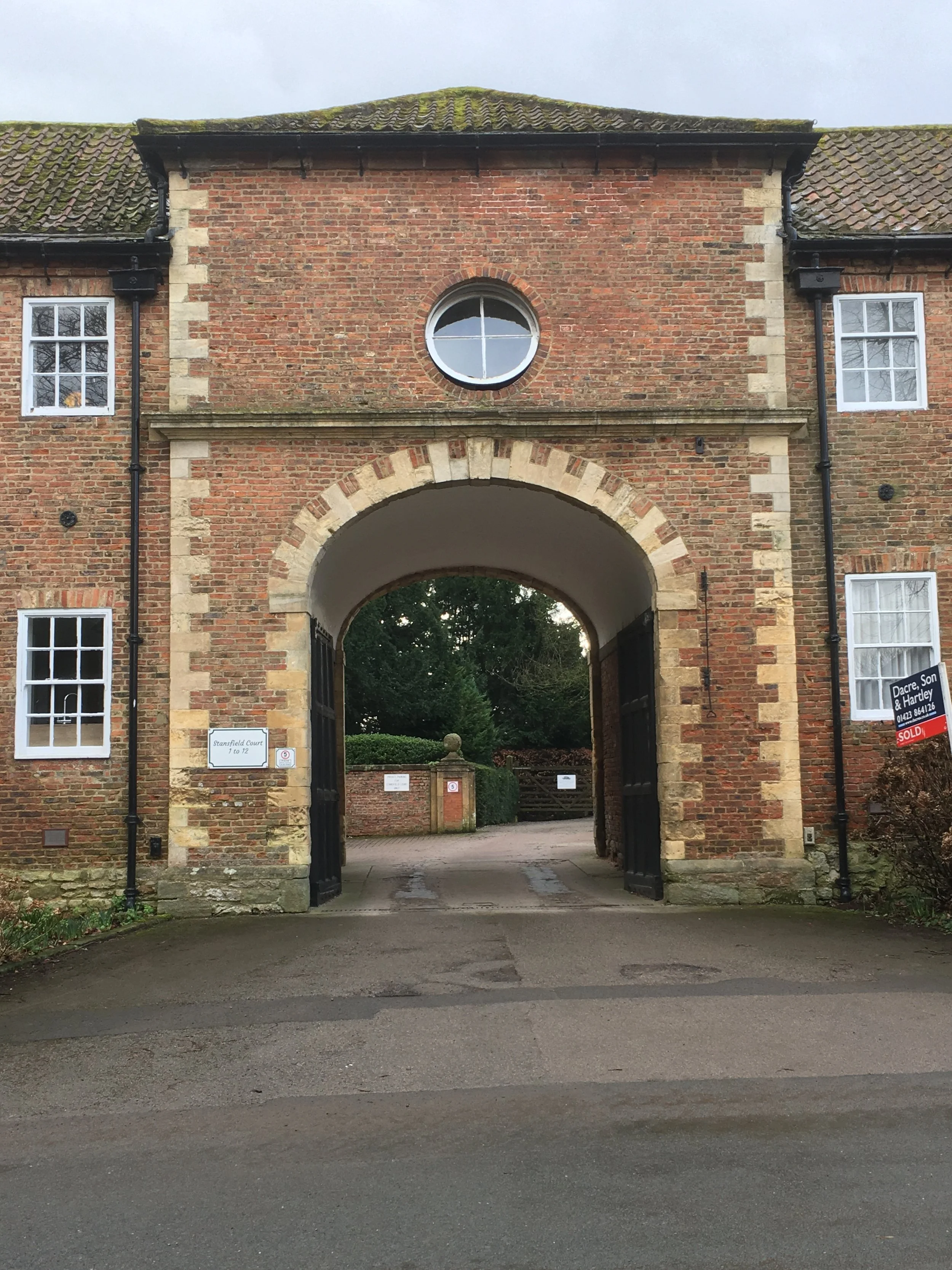 A brick building with a large archway entrance, four windows, and a round window above the archway, surrounded by greenery.