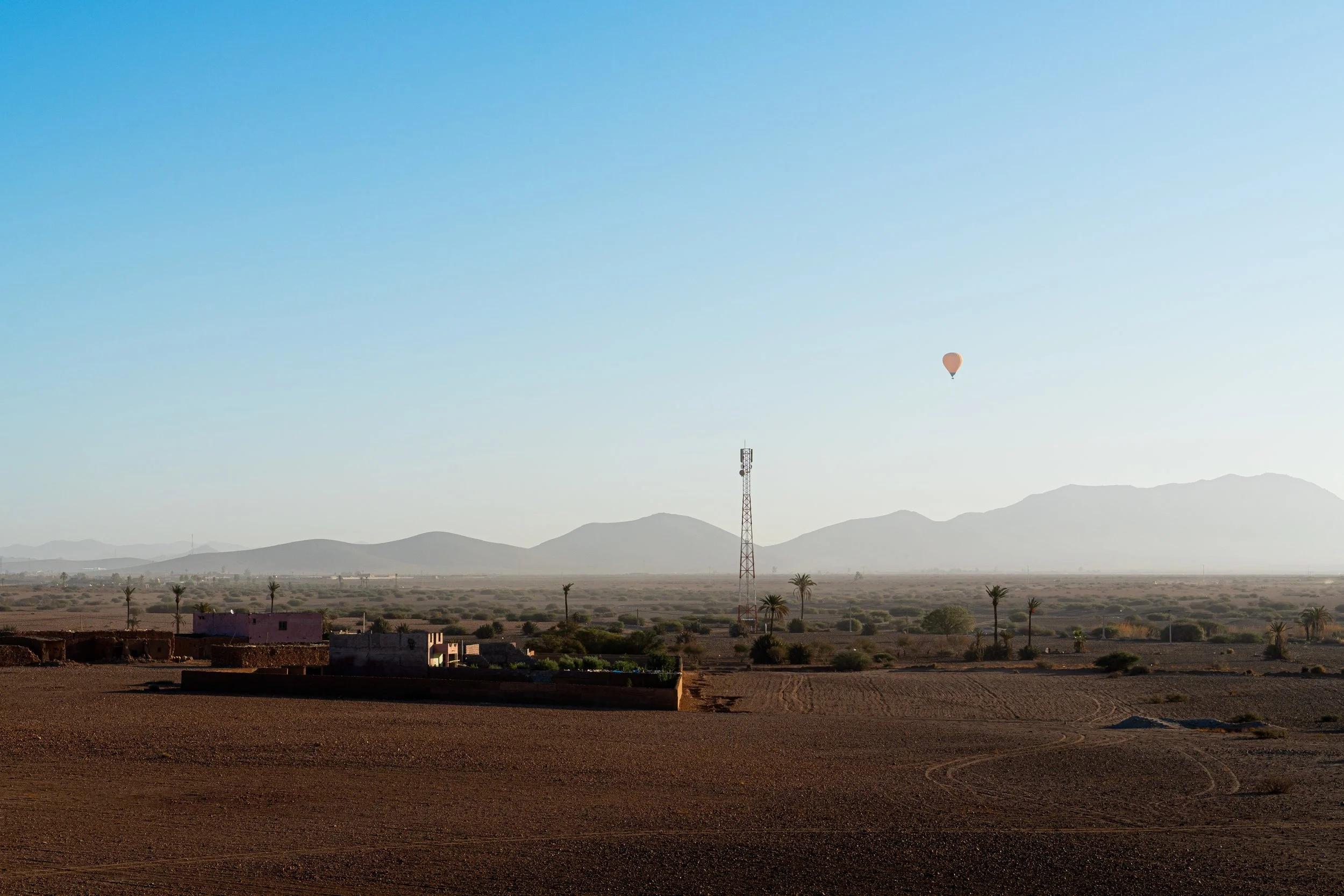 A desert landscape with a few small buildings, palm trees, and a communications tower in the distance. A hot air balloon floats in the clear blue sky above.