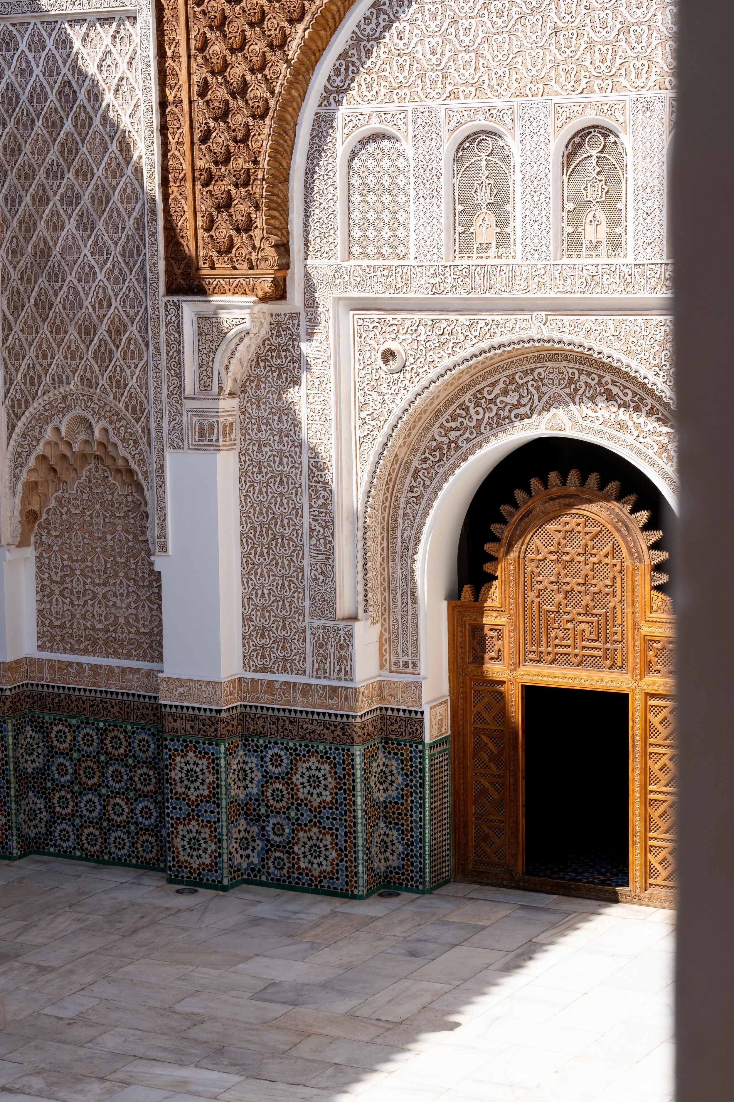 Decorative interior wall with intricate white carvings, arched niches, colorful tile work, and a wooden door with geometric patterns, part of traditional Islamic architecture.