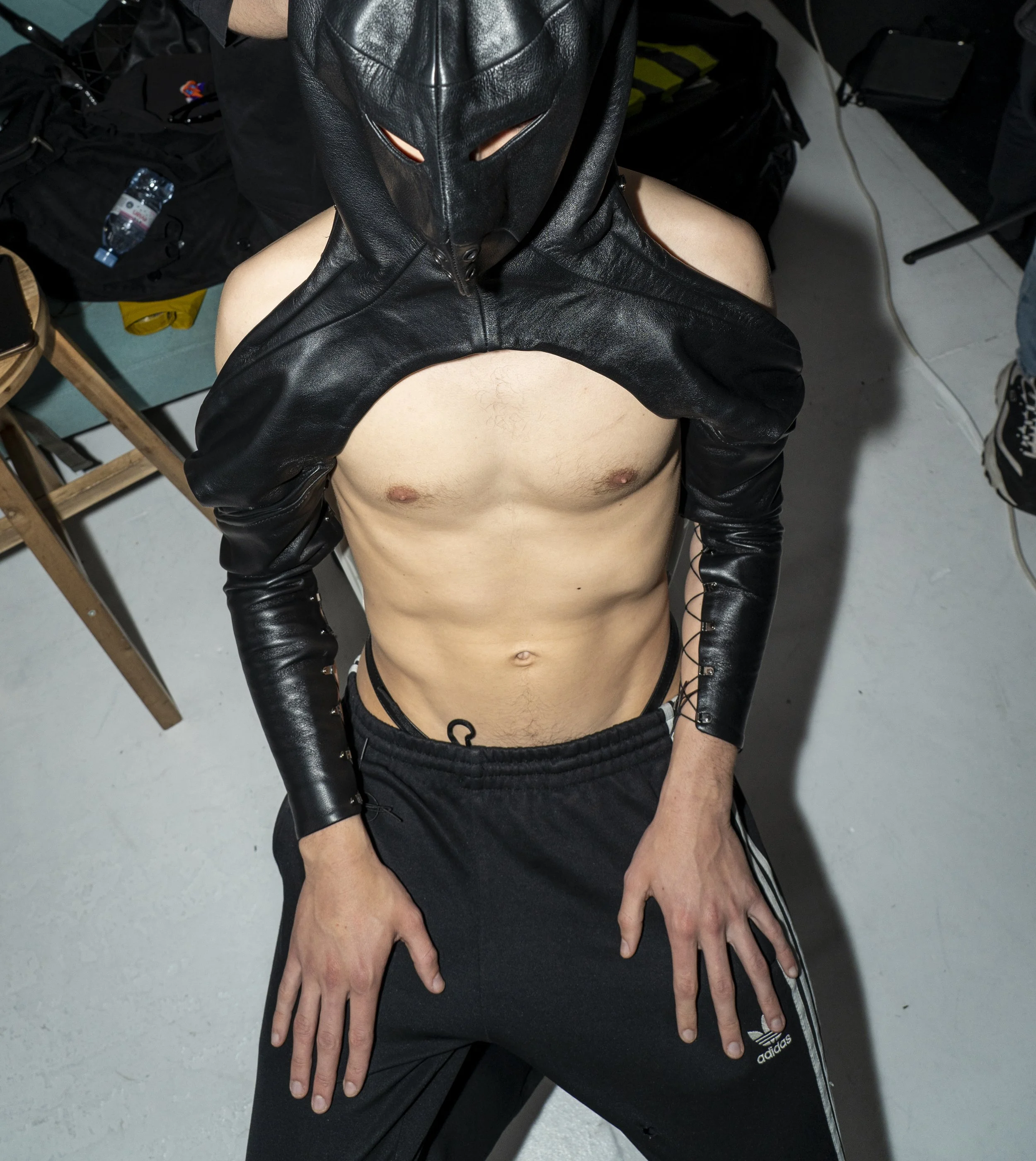 A shirtless man wearing black Adidas sweatpants and a black leather jacket with cut-out details, and a black mask with horns, standing indoors.