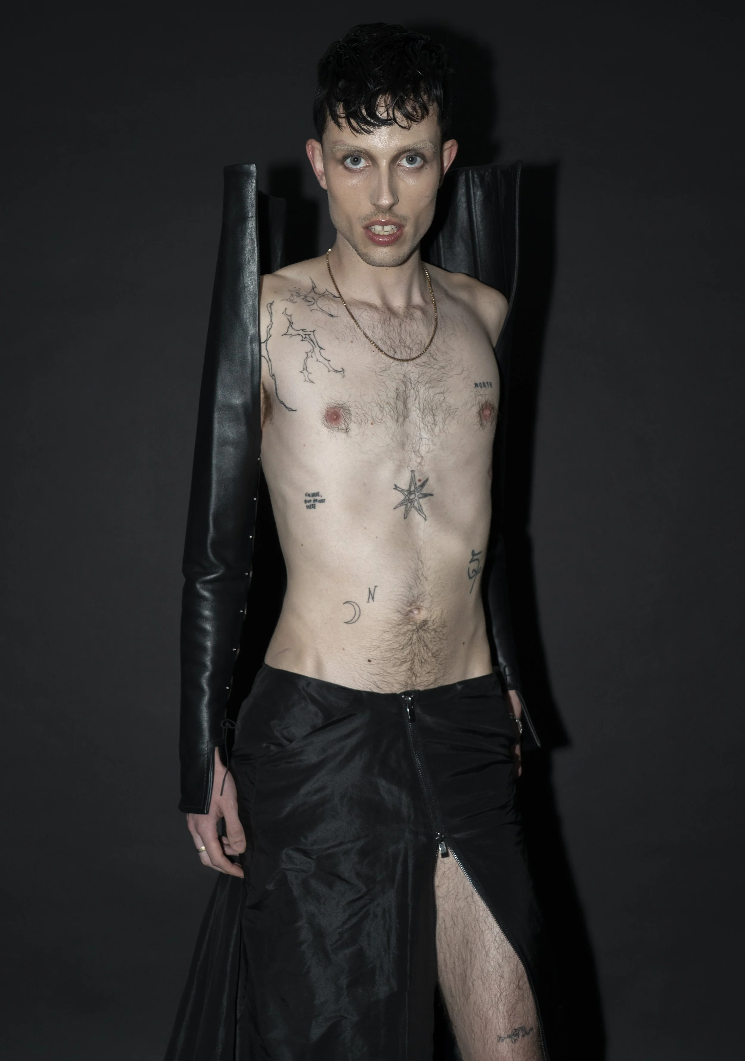 A young man with tattoos and short dark hair posing shirtless against a dark background, wearing black leather pants and a long black leather coat hanging off his shoulders.