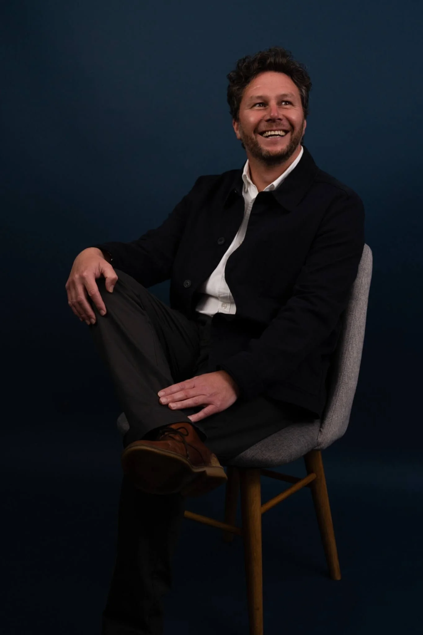 Paul Cartwright, Associate Director sitting on a grey chair against a dark background, smiling and looking to his left, wearing a black jacket, white shirt, dark pants, and brown shoes.