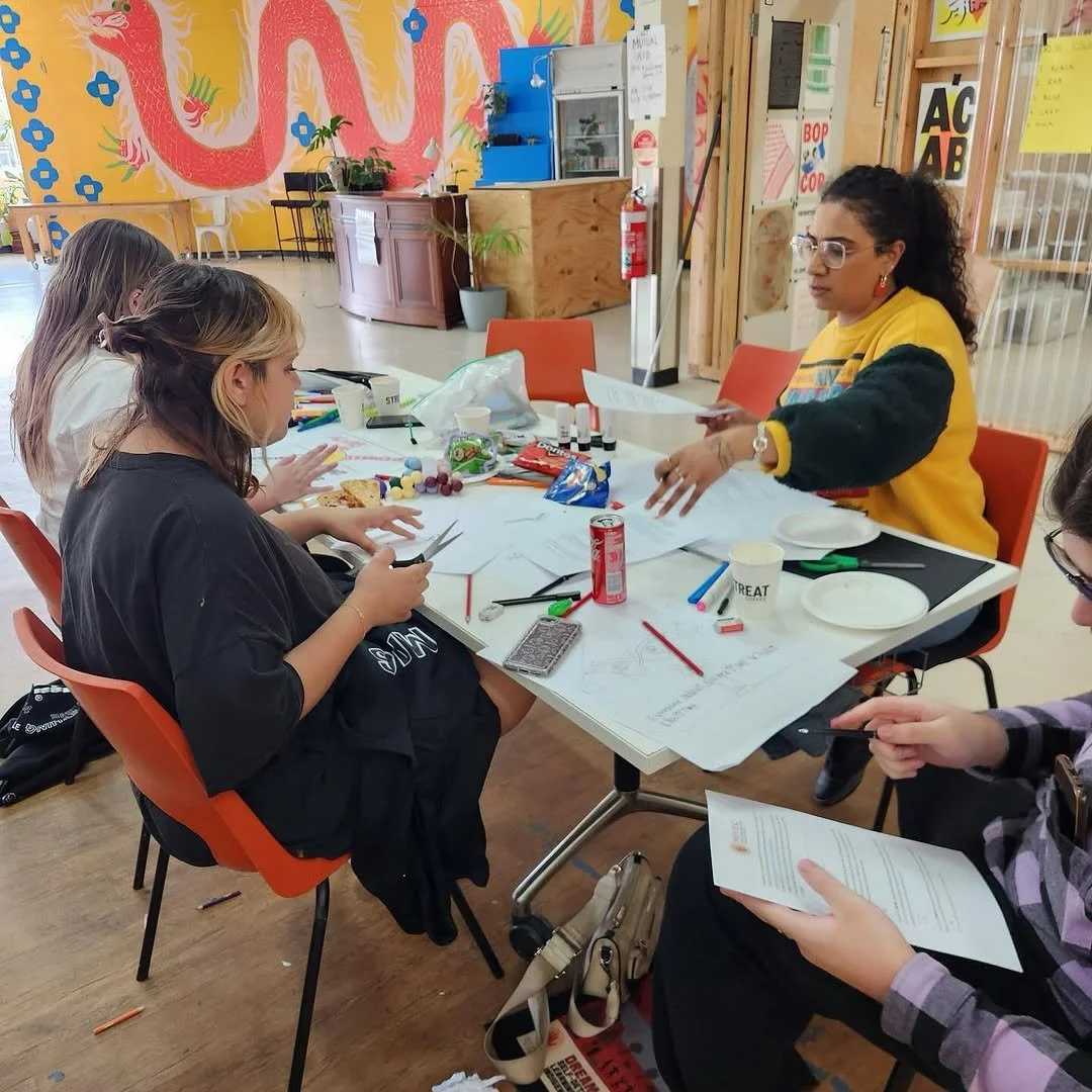 A group of young girls sitting around a table engaged in arts and crafts activities, with supplies such as papers, markers, glue, and scissors visible on the table, and a woman instructing them.
